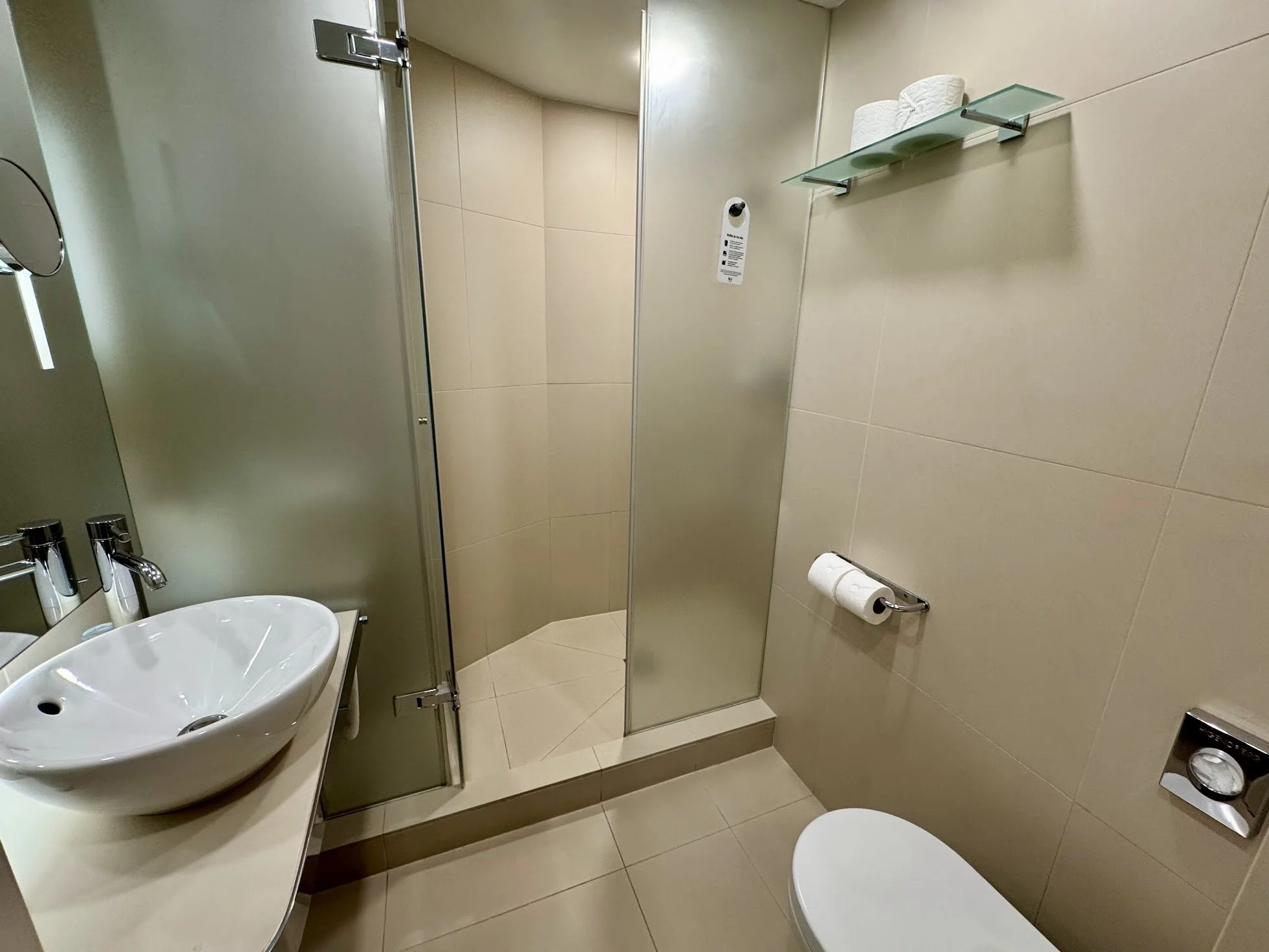 A small bathroom with a white sink and a circular mirror, beige tiled walls, a glass shower stall with a door, a toilet with a toilet paper roll, and a wall-mounted shelf with two rolls of toilet paper. There is also a soap dispenser on the sink and a small, black electronic device on the floor.