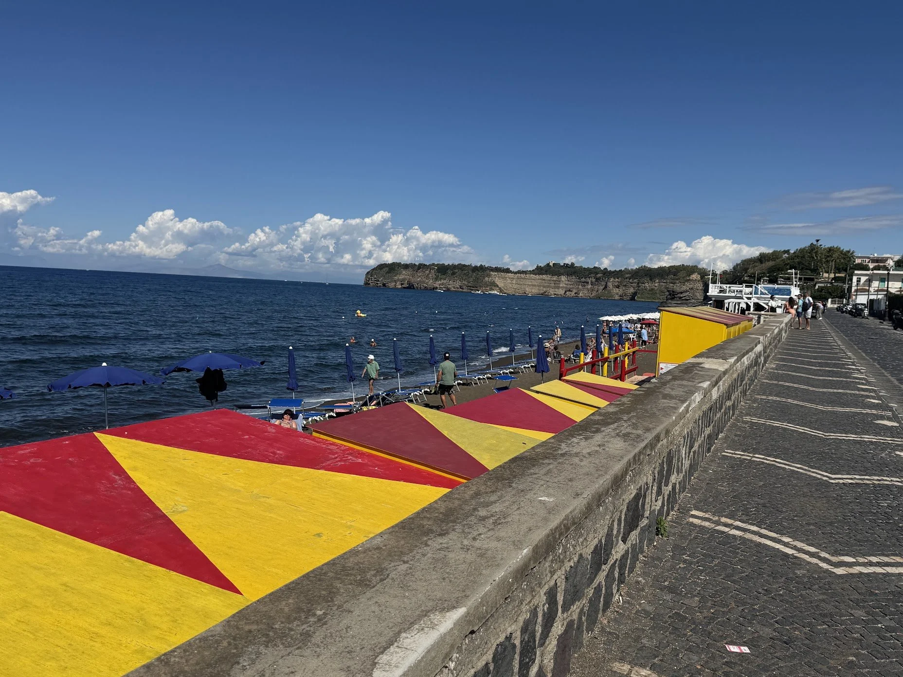 View of a beach with colorful yellow and red umbrellas, a walking promenade with people, and the ocean with cliffs and clouds in the background.