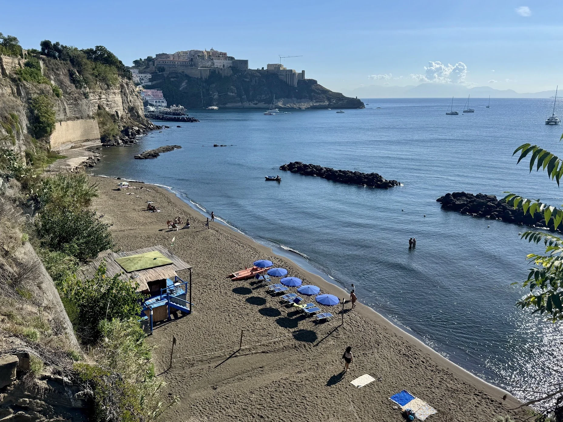 A beach with sand, blue umbrellas, and lounge chairs, bordered by a rocky cliff and vegetation on the left. Several people are on the beach and in the water. The calm sea has a few boats anchored, with a castle-like structure on a hill in the backgro