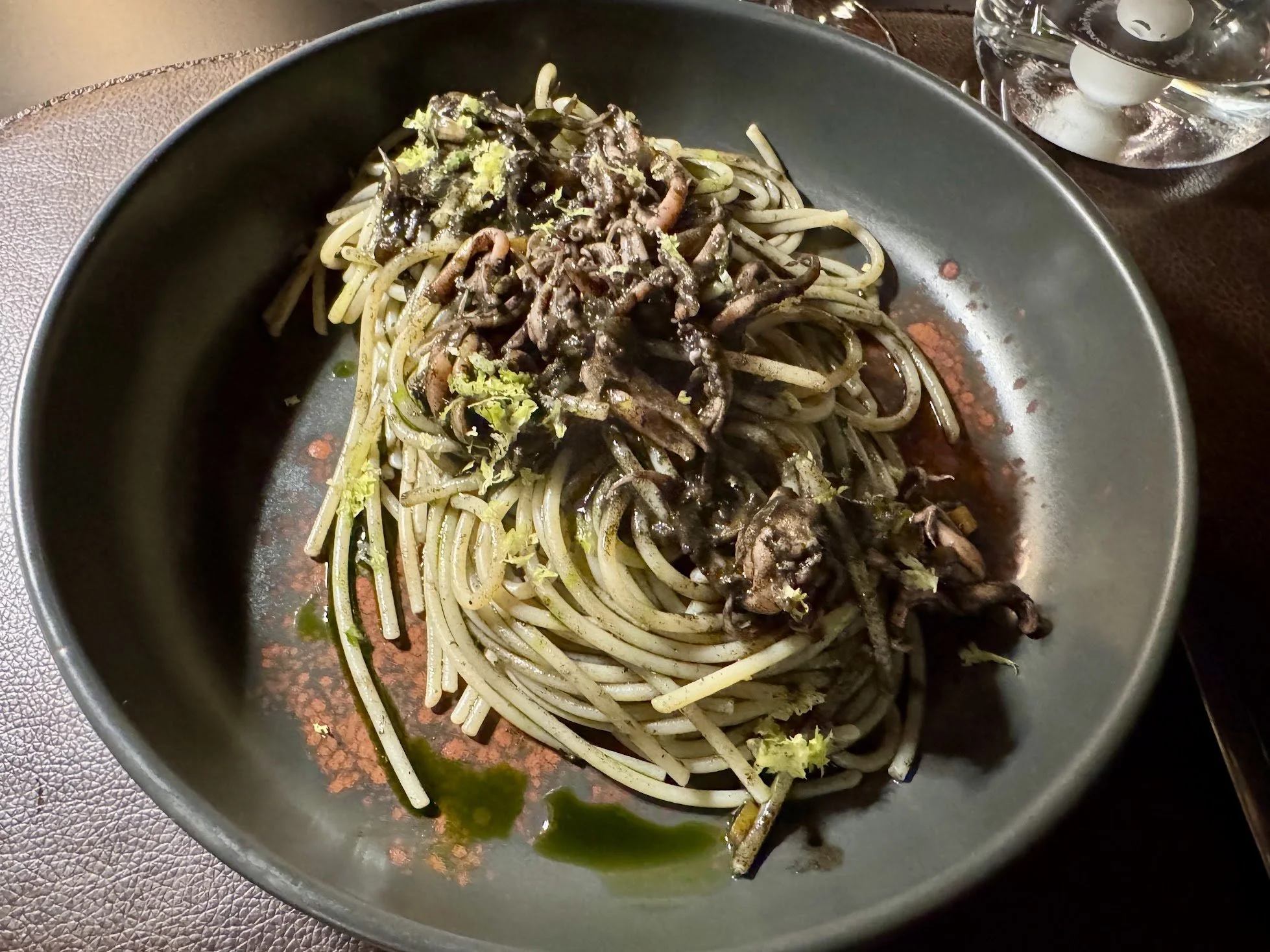 A black plate with spaghetti noodles topped with a dark sauce and some green lettuce pieces, with a glass of ice water nearby.