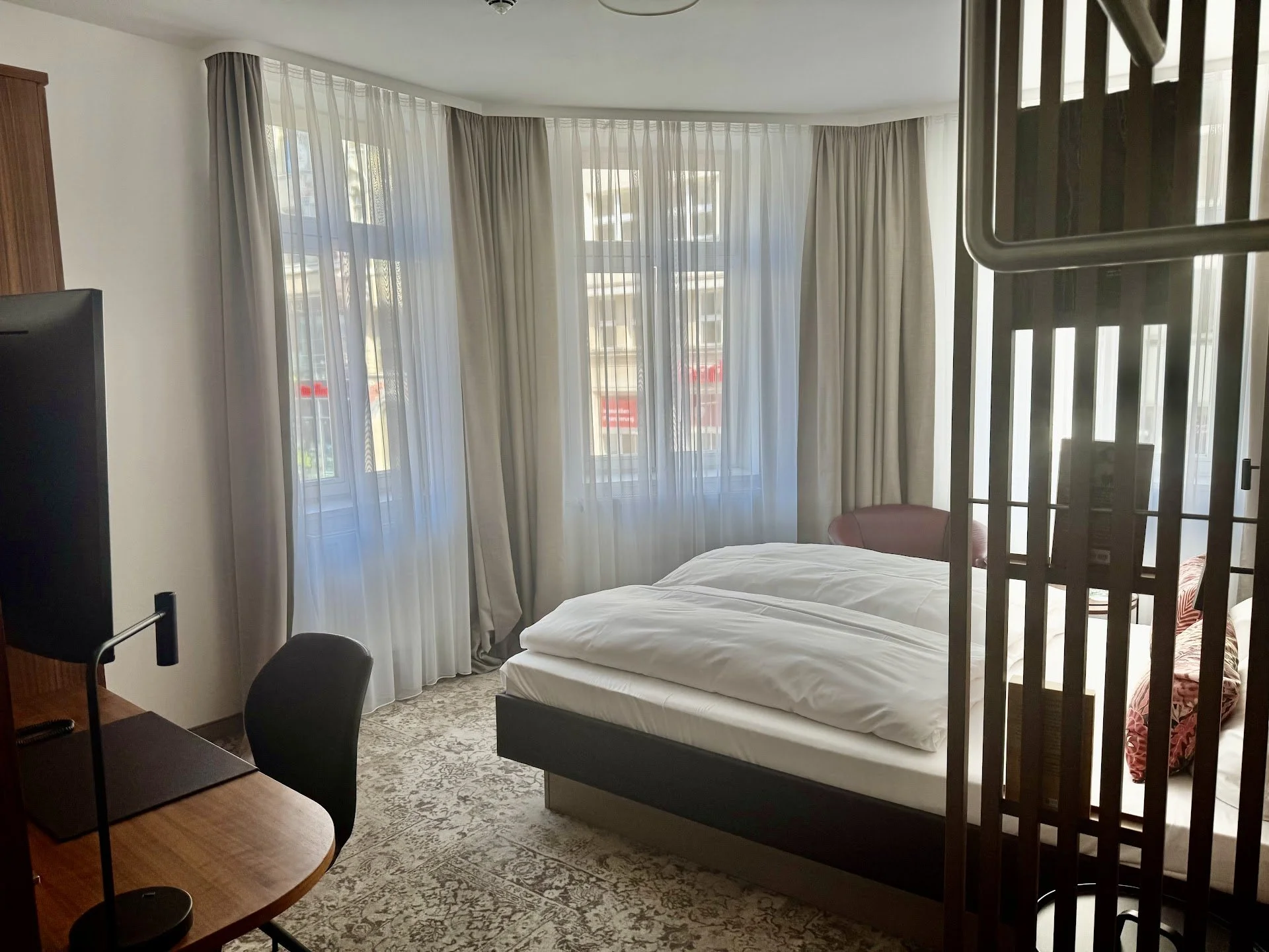 Hotel room with a bed near large windows covered with light curtains, a small round pink chair, a wooden desk with a chair, and a metal safety bar in the foreground.