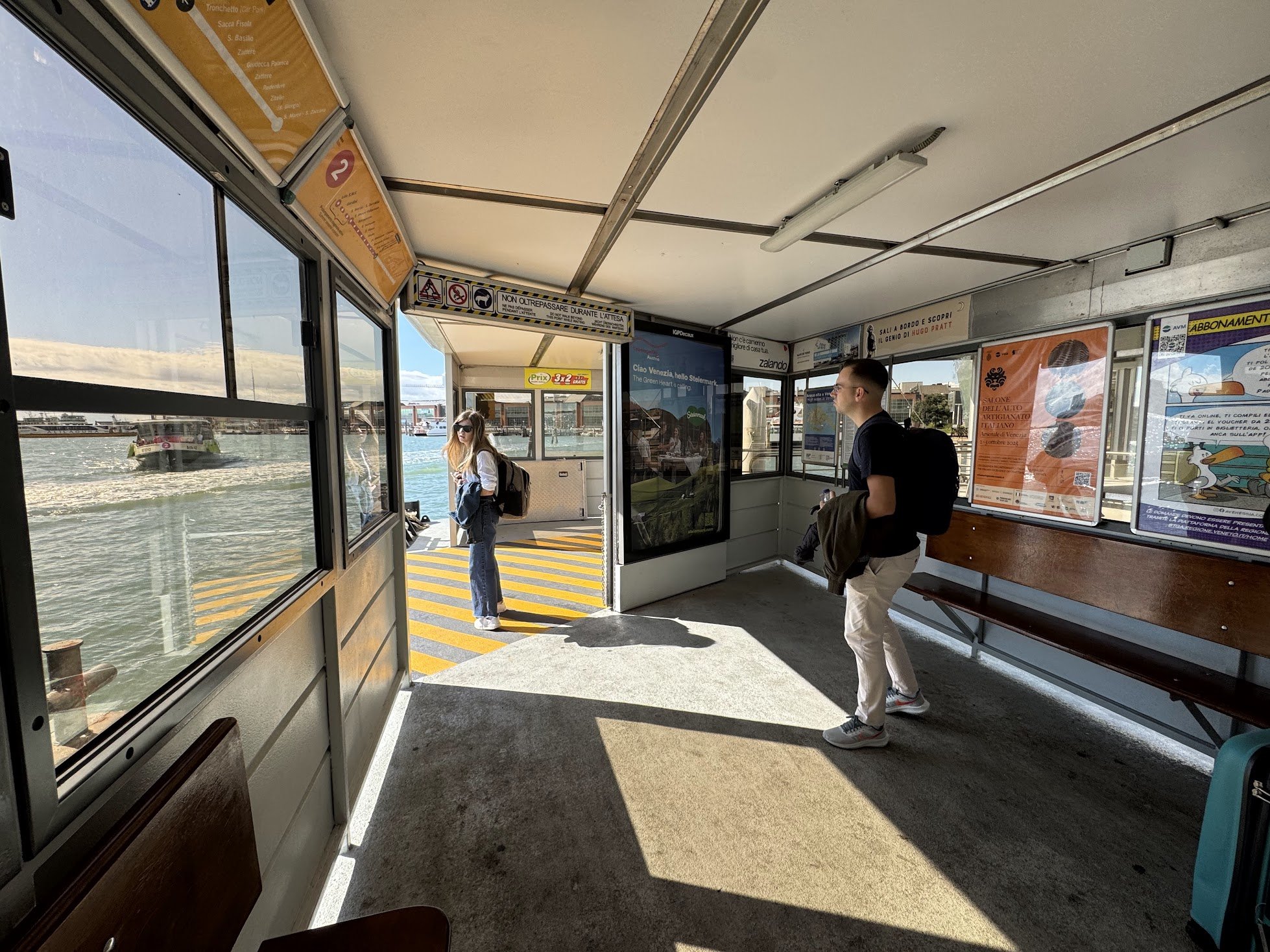 View inside a ferry terminal with two people waiting. One woman with sunglasses and a backpack is near the glass door, while a man in a dark jacket and white shoes is standing nearby. Outside, water and boats are visible.