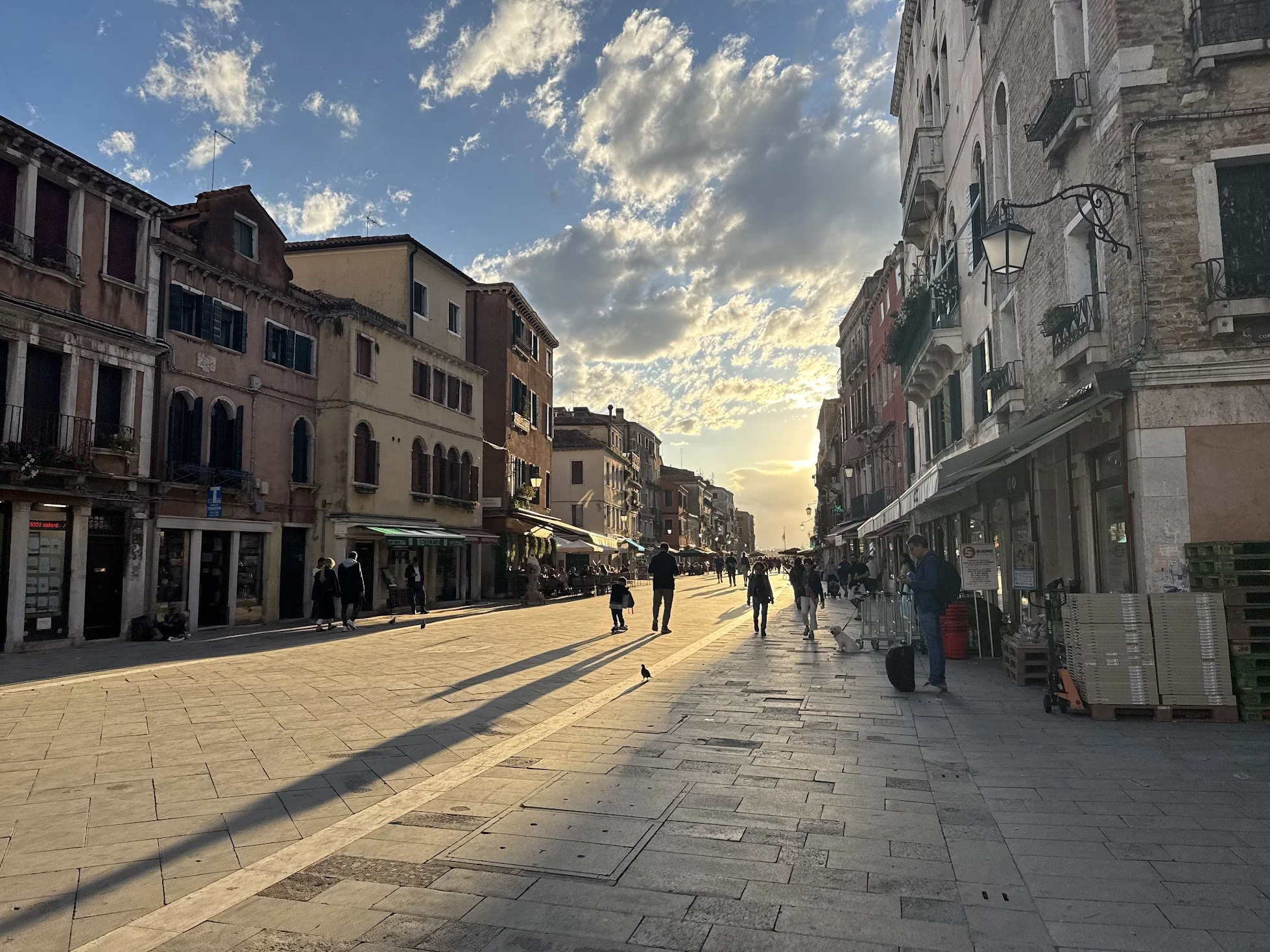 A street scene during sunset in a European city with people walking and shops on both sides. Buildings are multi-story with balconies, and the sky has scattered clouds.