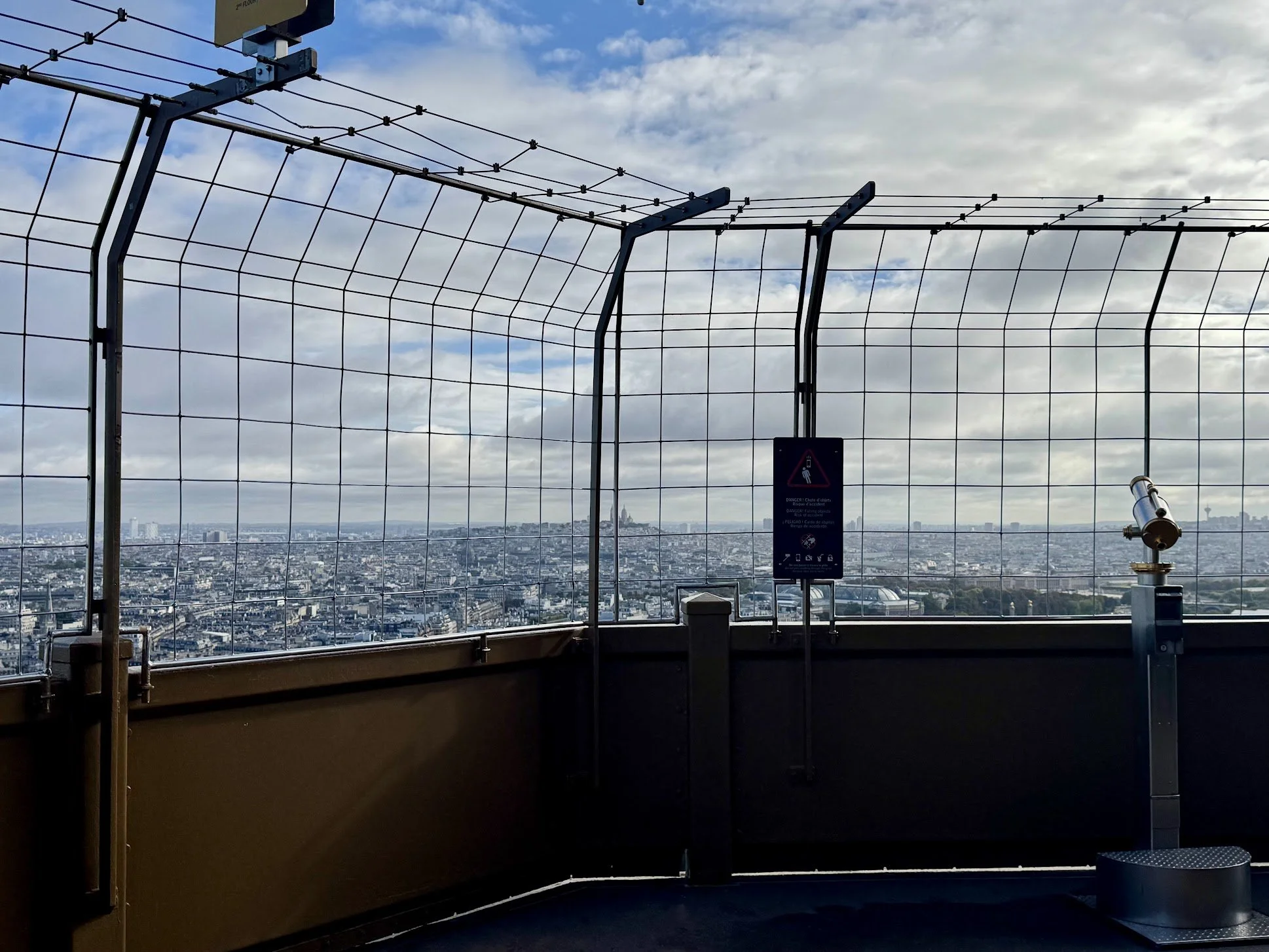 View from a tall building overlooking a cityscape through a metal safety fence, with cloudy sky overhead.