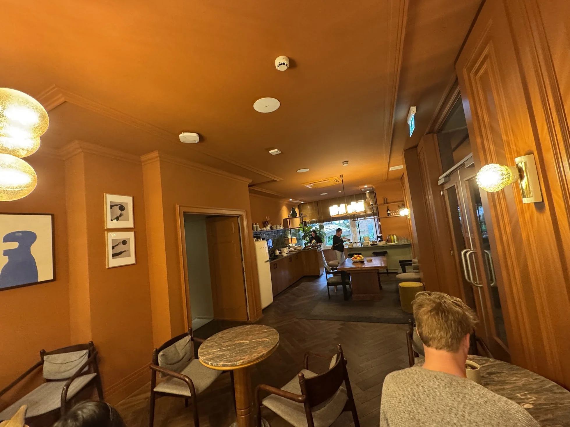 Interior of a cozy cafe or restaurant with warm yellow walls, wooden furniture, hanging light fixtures, and people seated at tables, with a visible kitchen area where staff are preparing food.