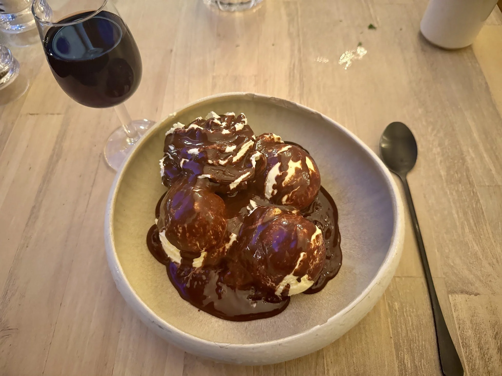 A bowl of ice cream topped with chocolate syrup, with a glass of red wine, a spoon, and water glasses on a wooden table.