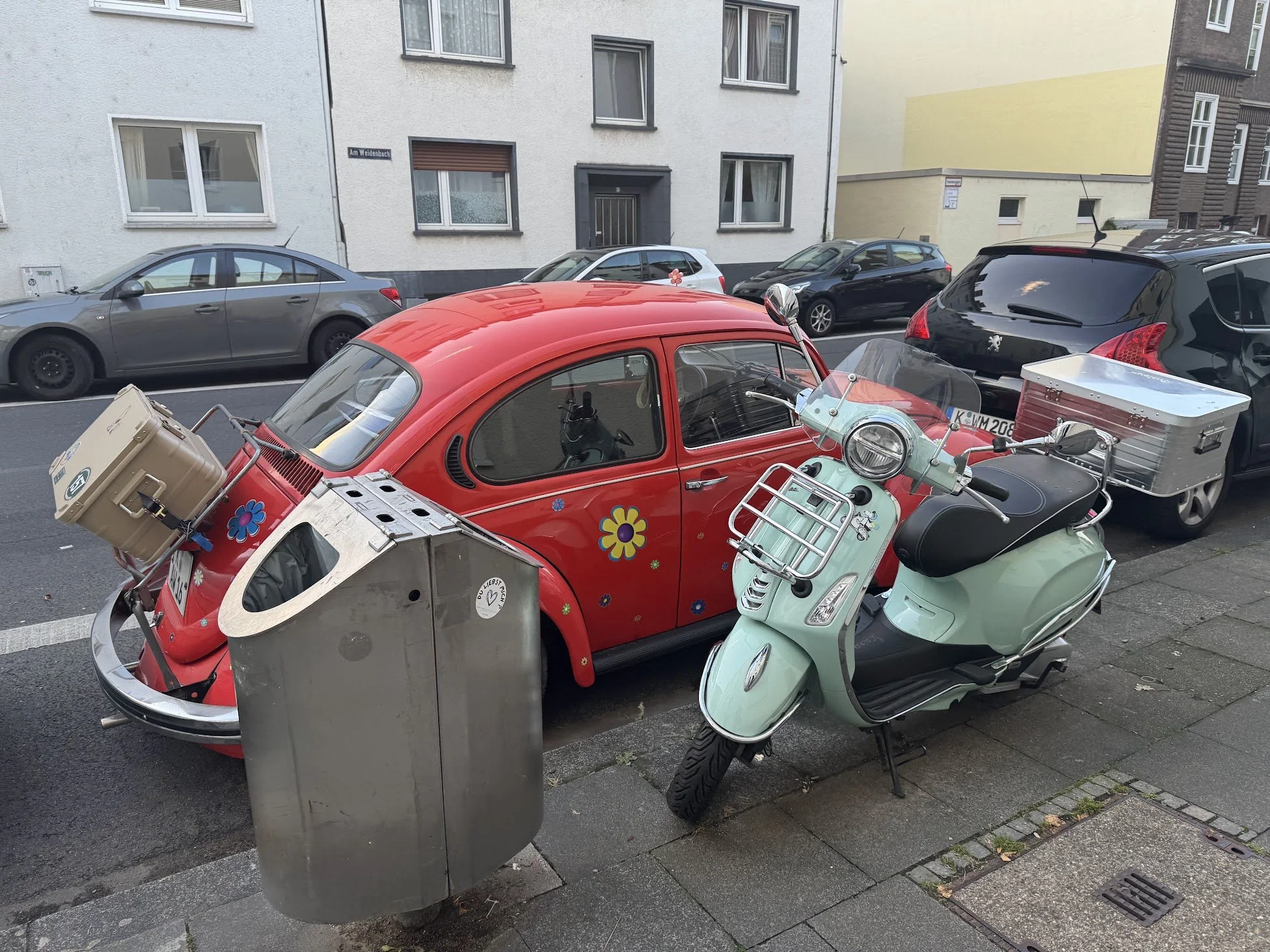 A red vintage Fiat 500 parked on the street beside several modern cars, with a grey trash bin and a mint green scooter nearby. Buildings with white and beige walls are visible in the background.