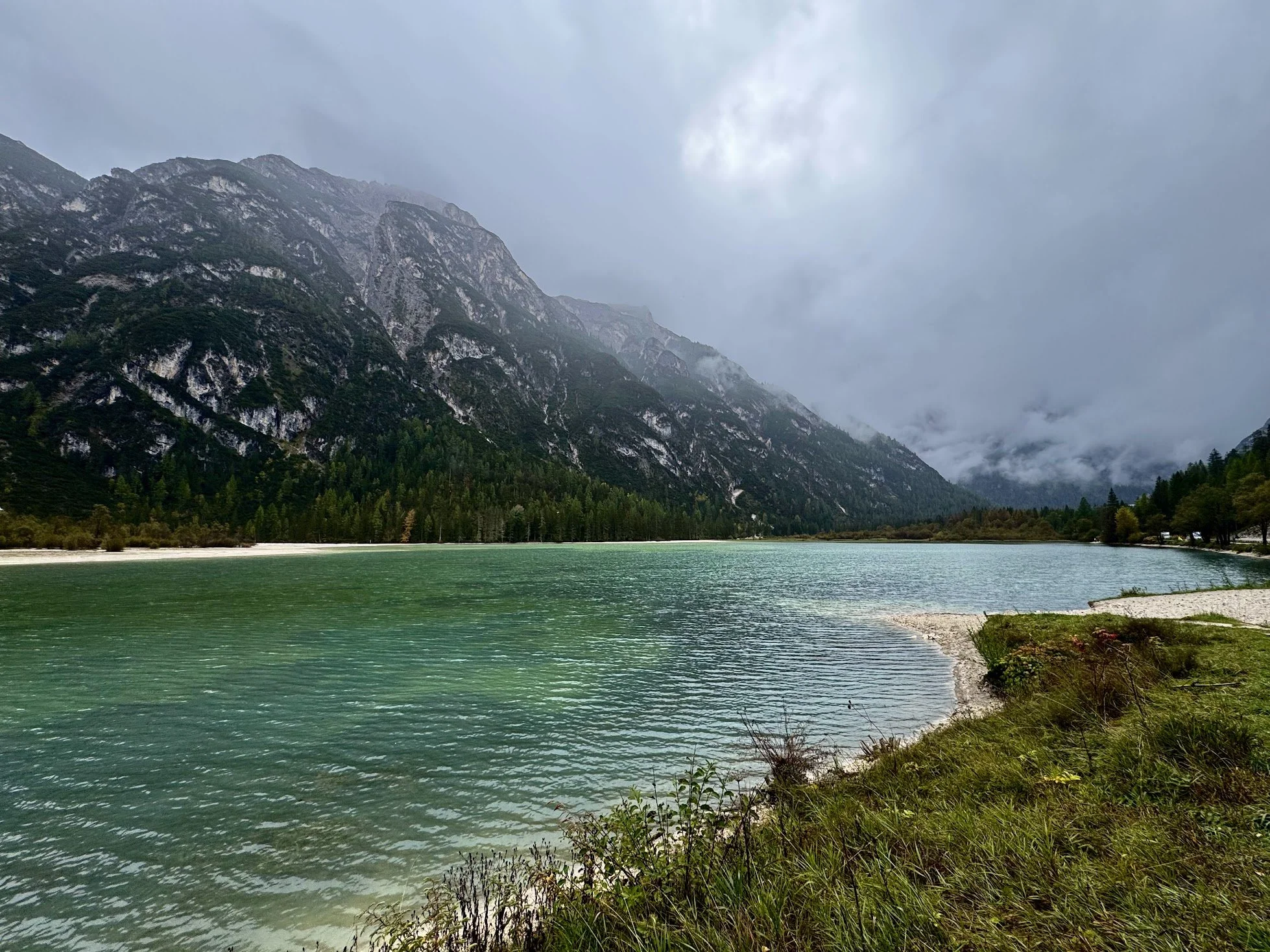 A mountain lake with green water surrounded by dense forest and high mountains partially covered in mist, under a cloudy sky.