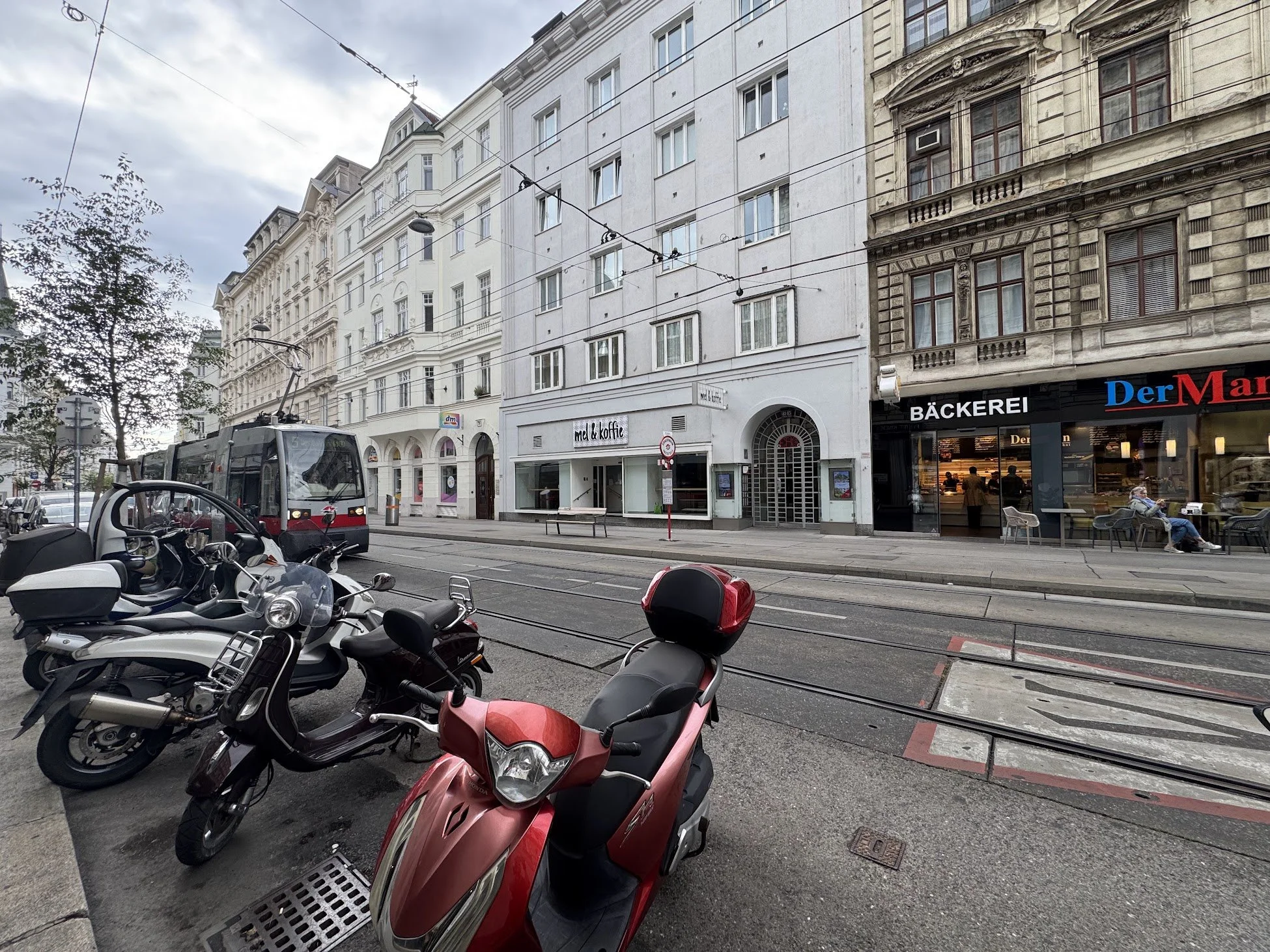 City street scene with parked motorcycles and scooters in the foreground, tram tracks, multi-story buildings with shops on the ground floor, and a tram in the background. Overhead tram wires and cloudy sky are visible.