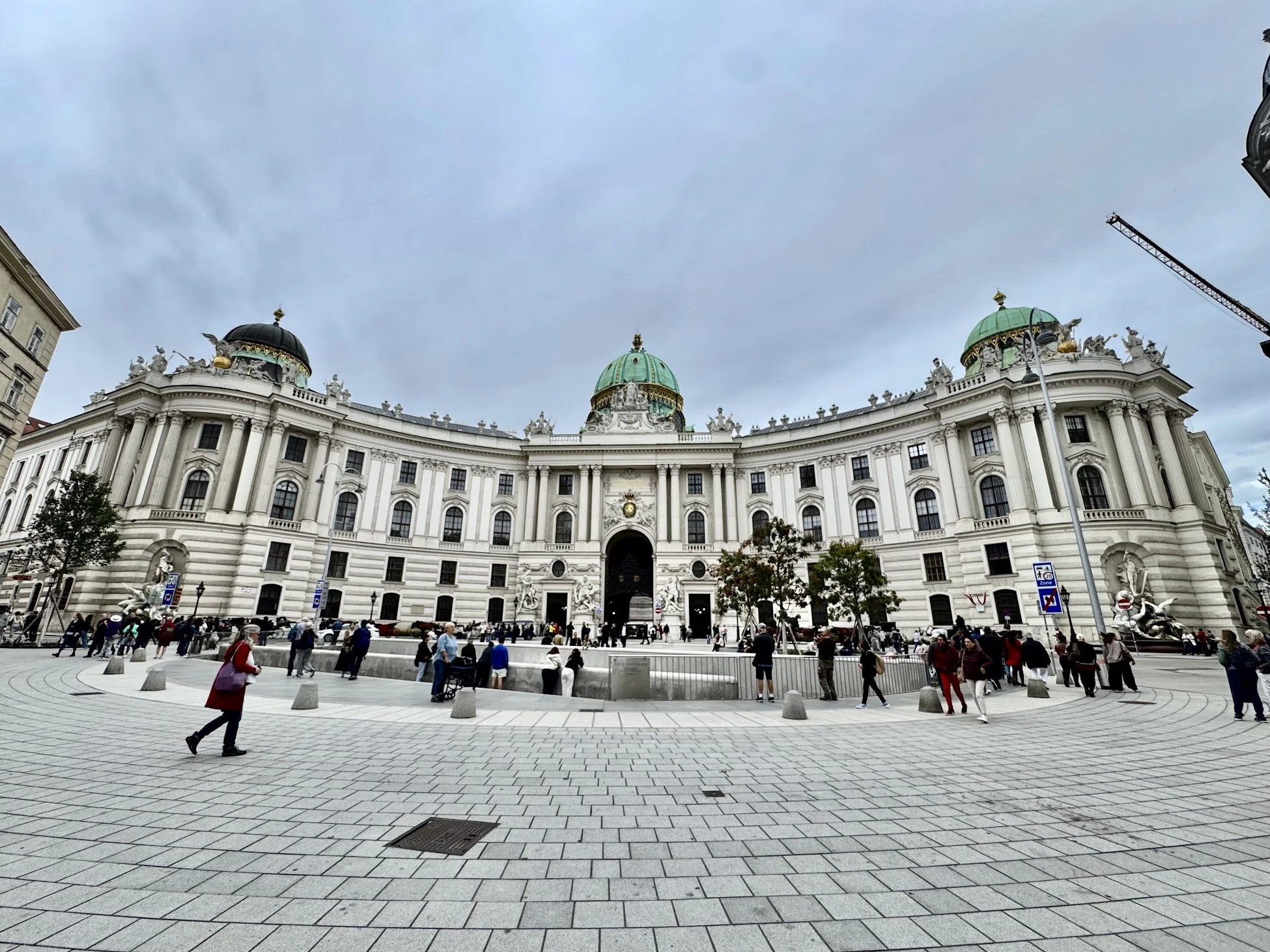 A large historic building with ornate architecture, green domed roofs, and decorative sculptures on top, situated in a busy square with pedestrians and trees in the foreground, and a construction crane visible in the background.