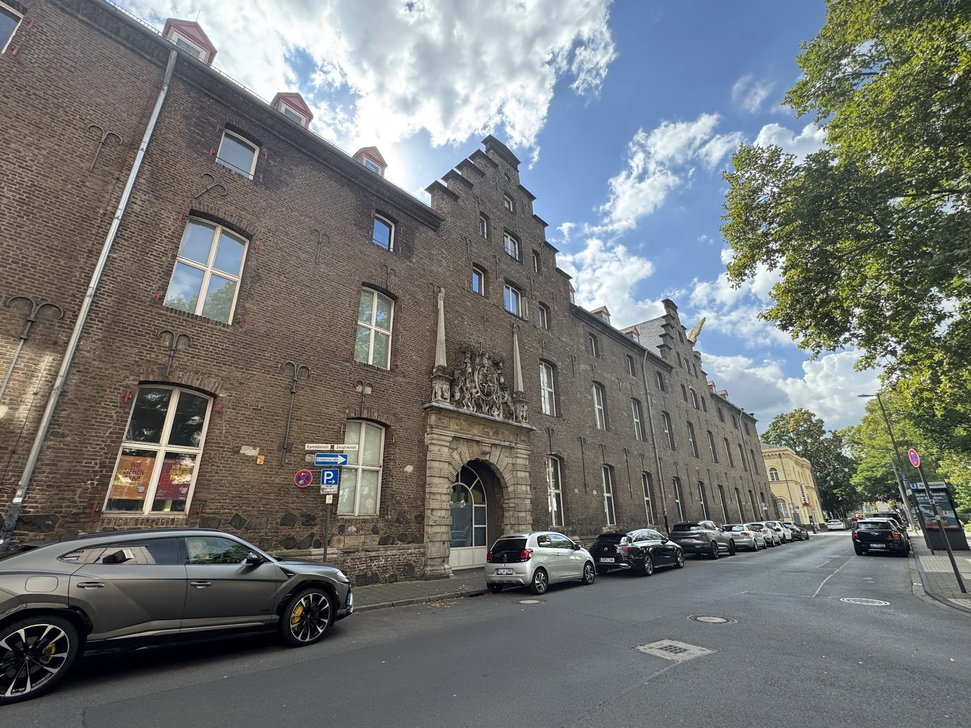 View of a historic brick building with decorative architectural details on a city street, with parked cars and a sidewalk, under a partly cloudy sky and some green trees.