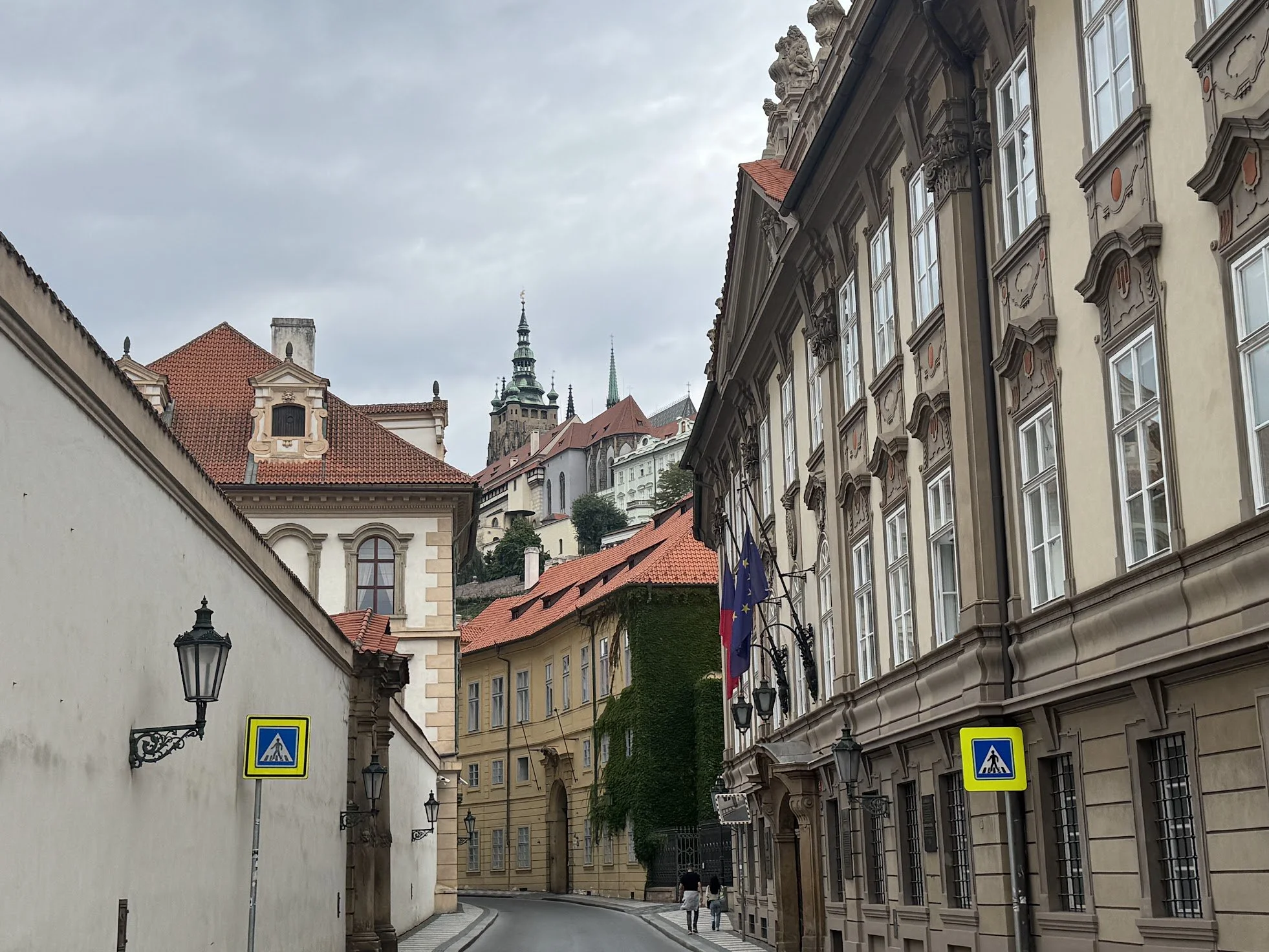 A narrow European street with historical buildings, two people walking, flags hanging outside a building, overcast sky.