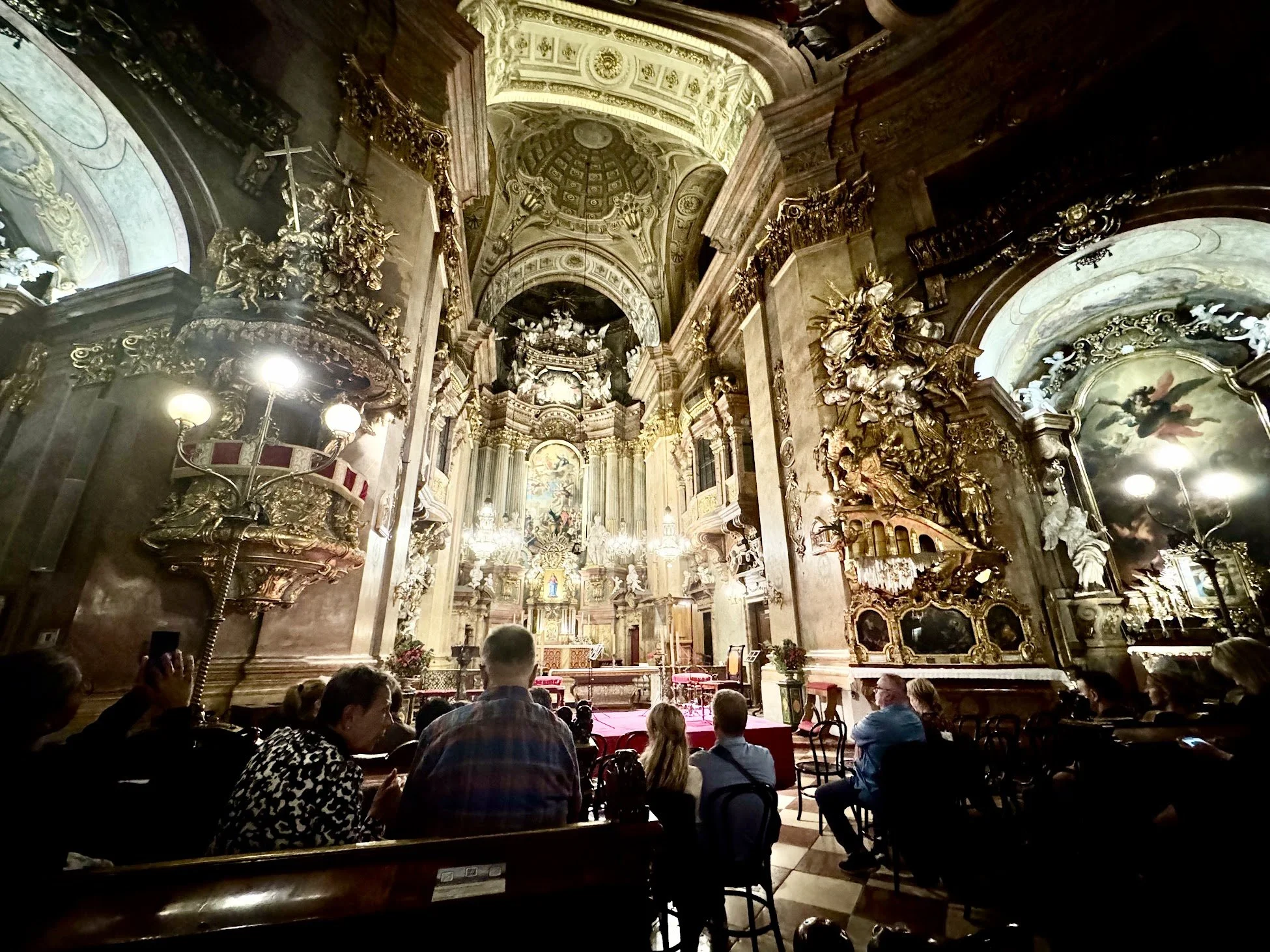Inside a richly decorated church or cathedral with gold accents, ornate sculptures, and religious paintings, gathered with people seated facing the altar.