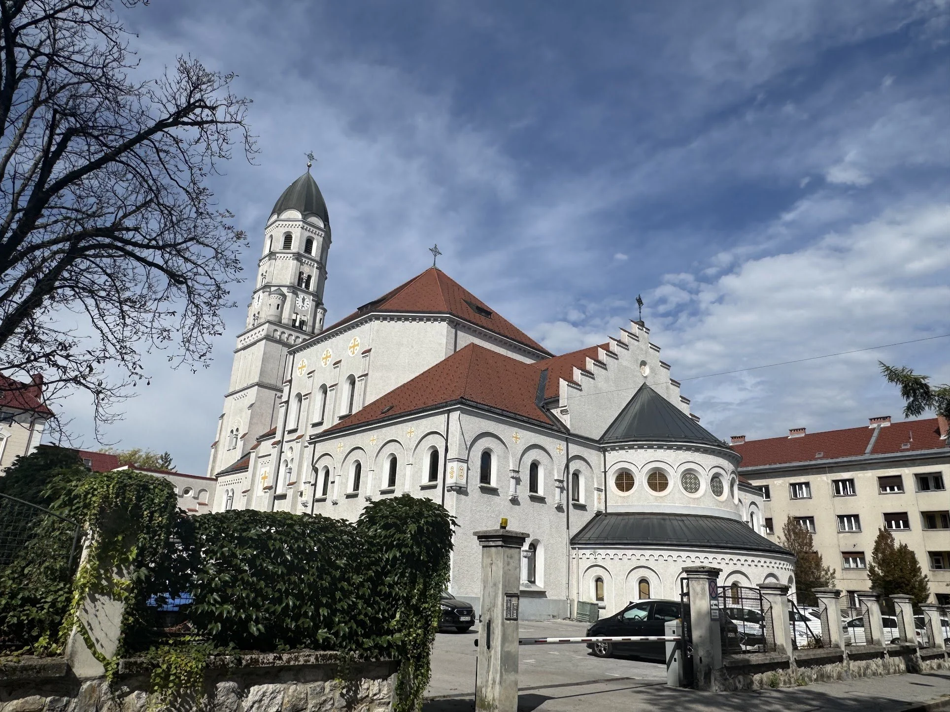 A white church with a tall steeple featuring a clock and weather vane, red-tiled roof, rounded windows, and decorative cross symbols, set against a partly cloudy sky and surrounded by trees and parked cars.