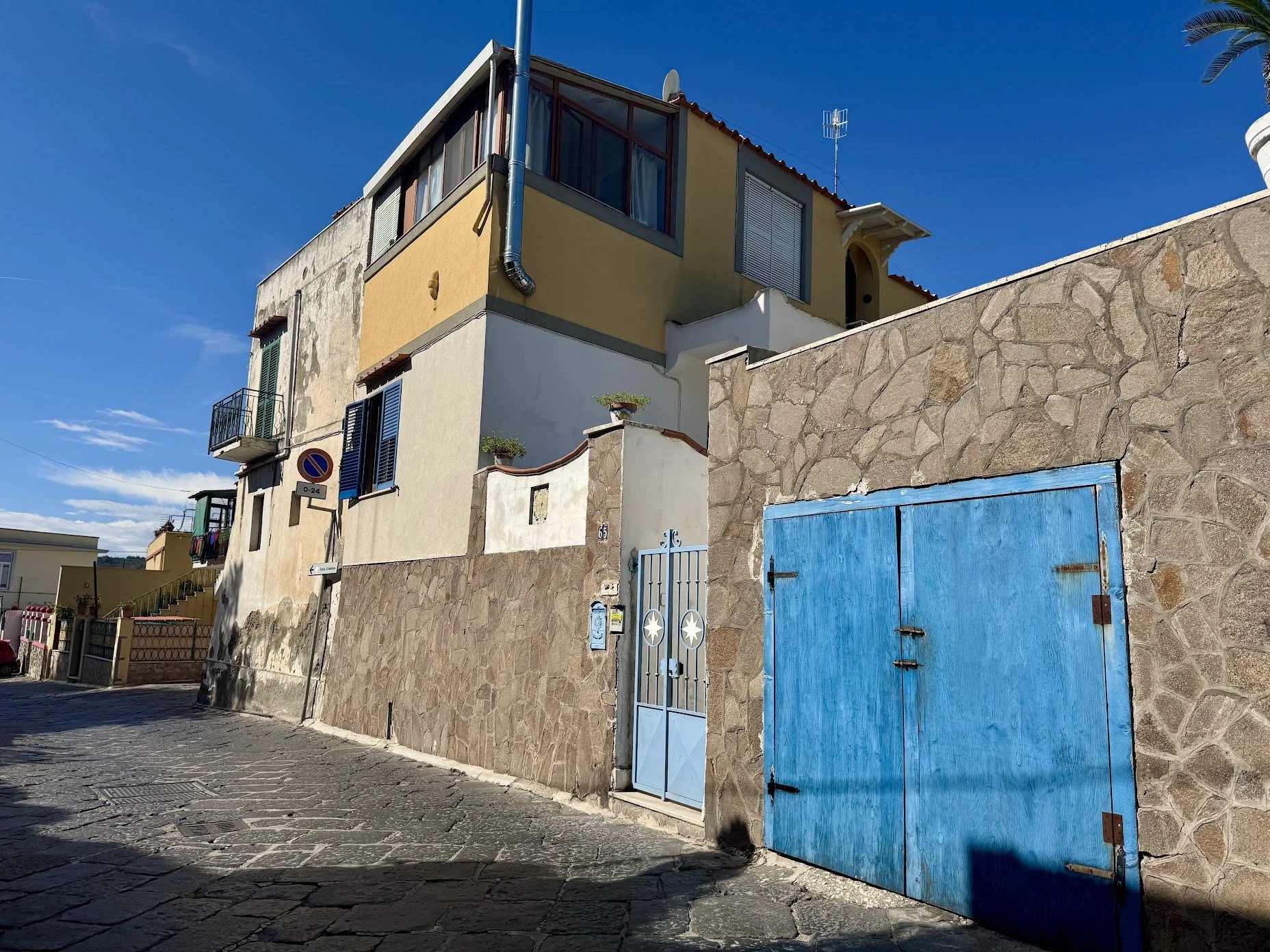 A street scene showing a multi-story building with a stone wall and a closed blue gate and door, blue sky, and some greenery.
