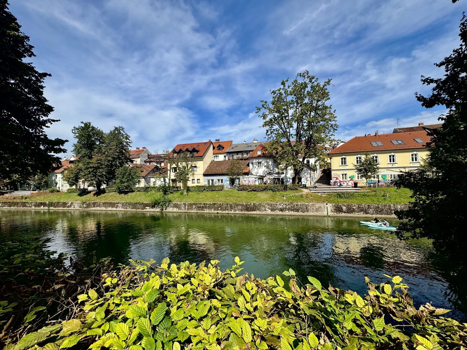 A scenic view of a river with houses and trees along the banks, and a boat with two people paddling on the water. The sky is partly cloudy with some blue sky visible.
