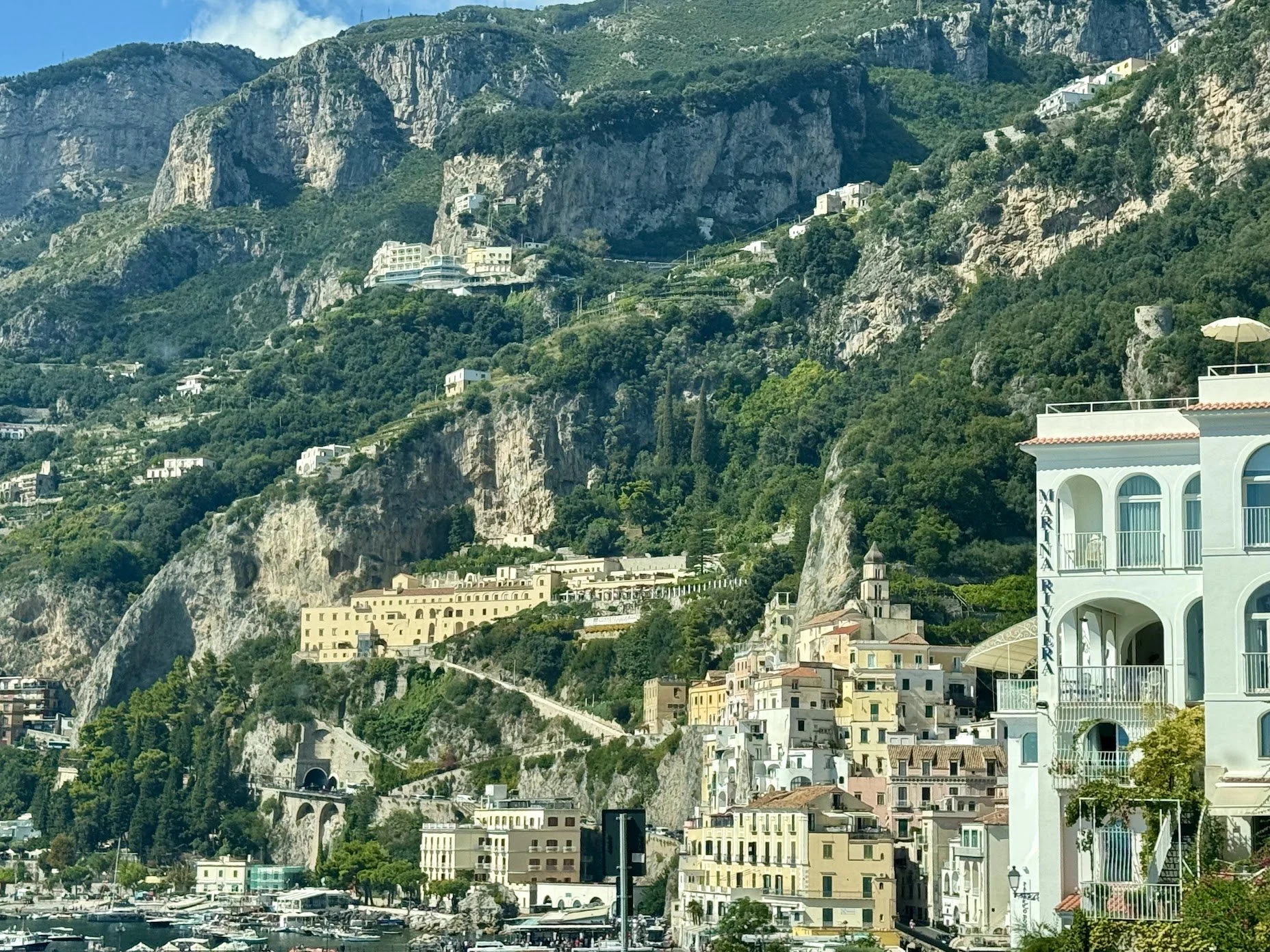 Cliffside view of a coastal town with colorful buildings and lush green vegetation, hillside tunnels, and boats at the waterfront.
