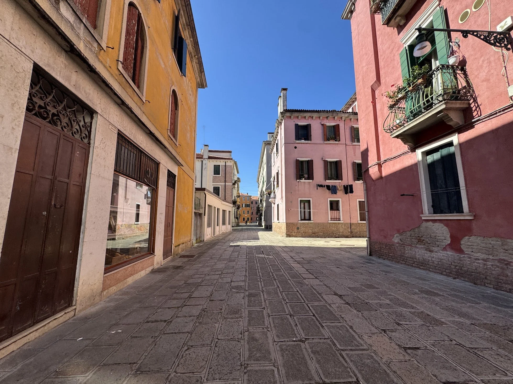 A narrow cobblestone street in a European city with colorful buildings on either side, blue sky overhead, and a person walking in the distance.