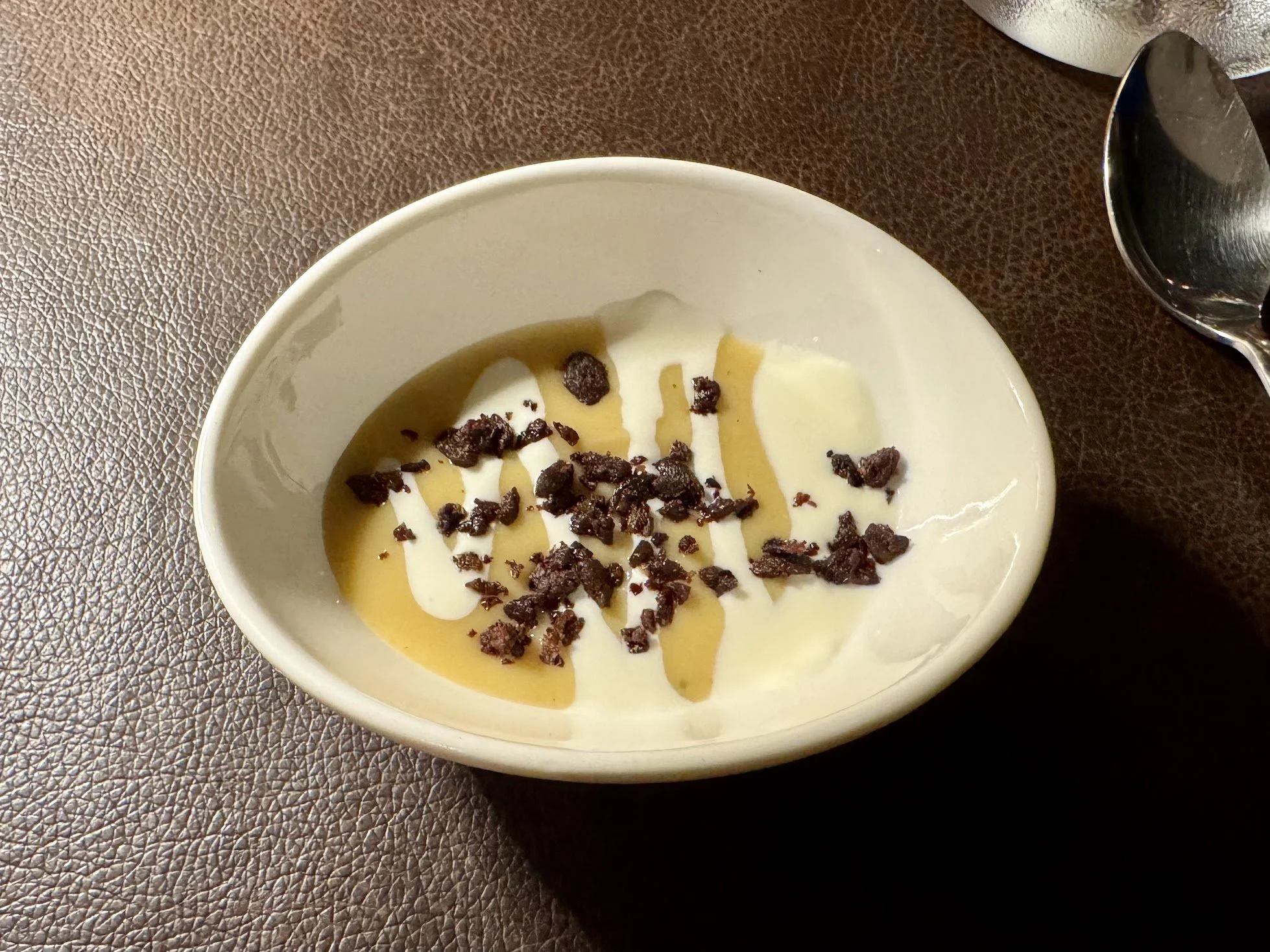A bowl of dessert with white yogurt or cream, caramel sauce drizzled on top, and sprinkled crushed chocolate or cookie crumbs, on a brown textured surface with a spoon nearby.