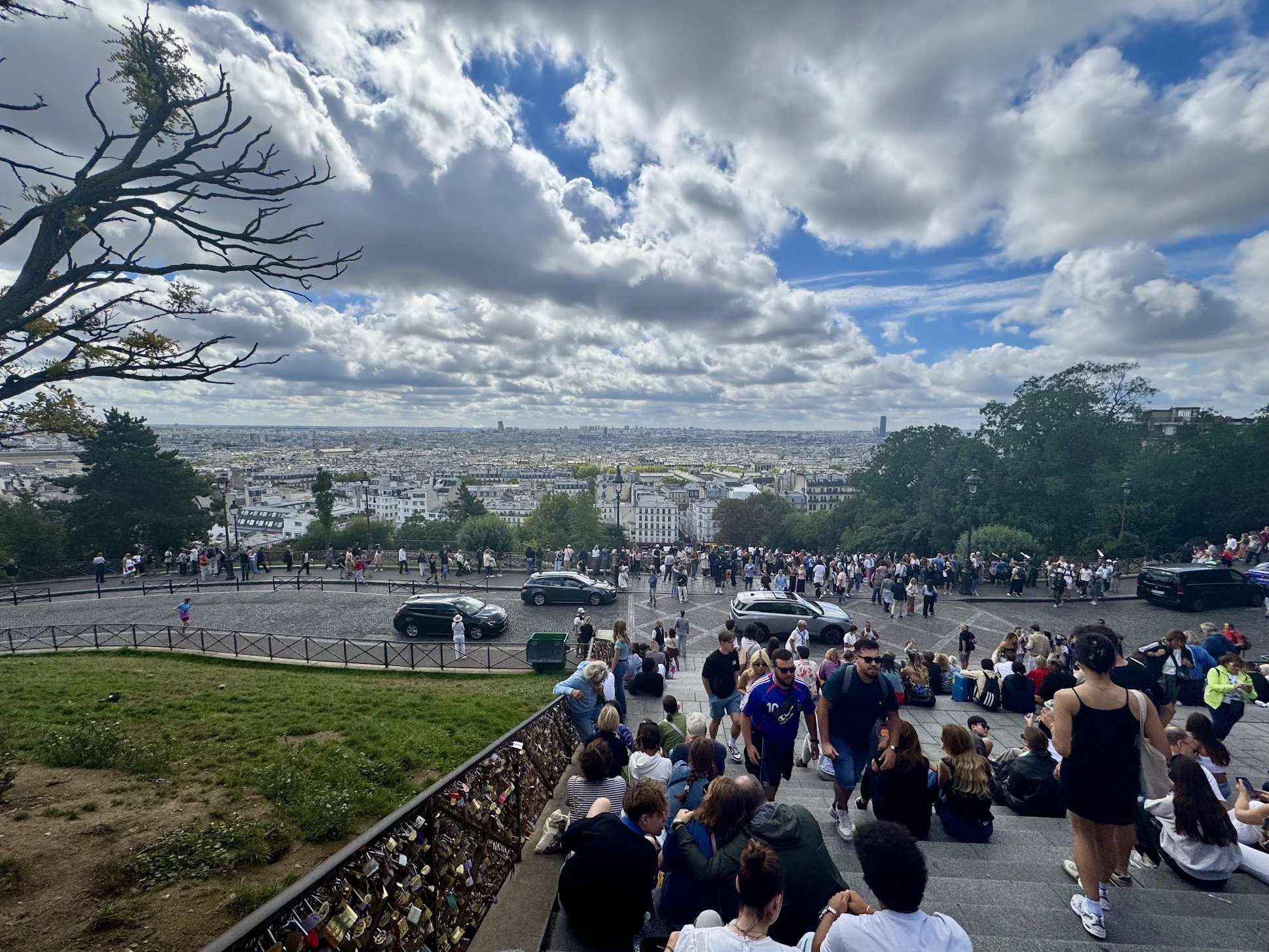 Crowd of tourists sitting and standing on a broad staircase, with a view of Paris cityscape under a partly cloudy sky in the background, and locks attached to the railing in the foreground.