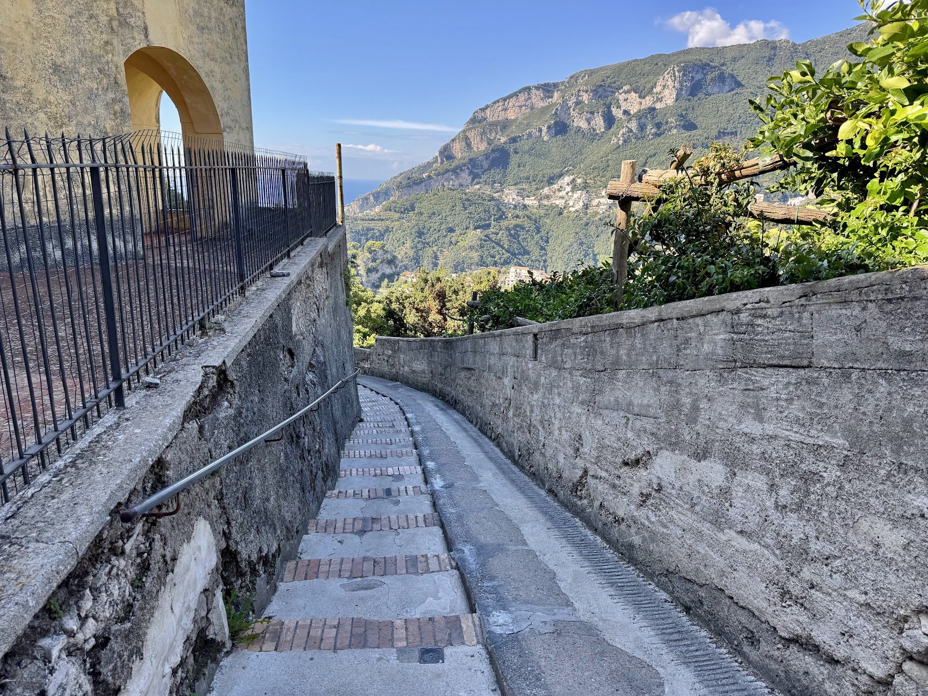 A narrow stone pathway with brick and concrete paving, bordered by stone walls, winding through a hillside town. To the left, there is a building with a yellowish wall and an arched window, with a black metal fence along the edge. On the right, there