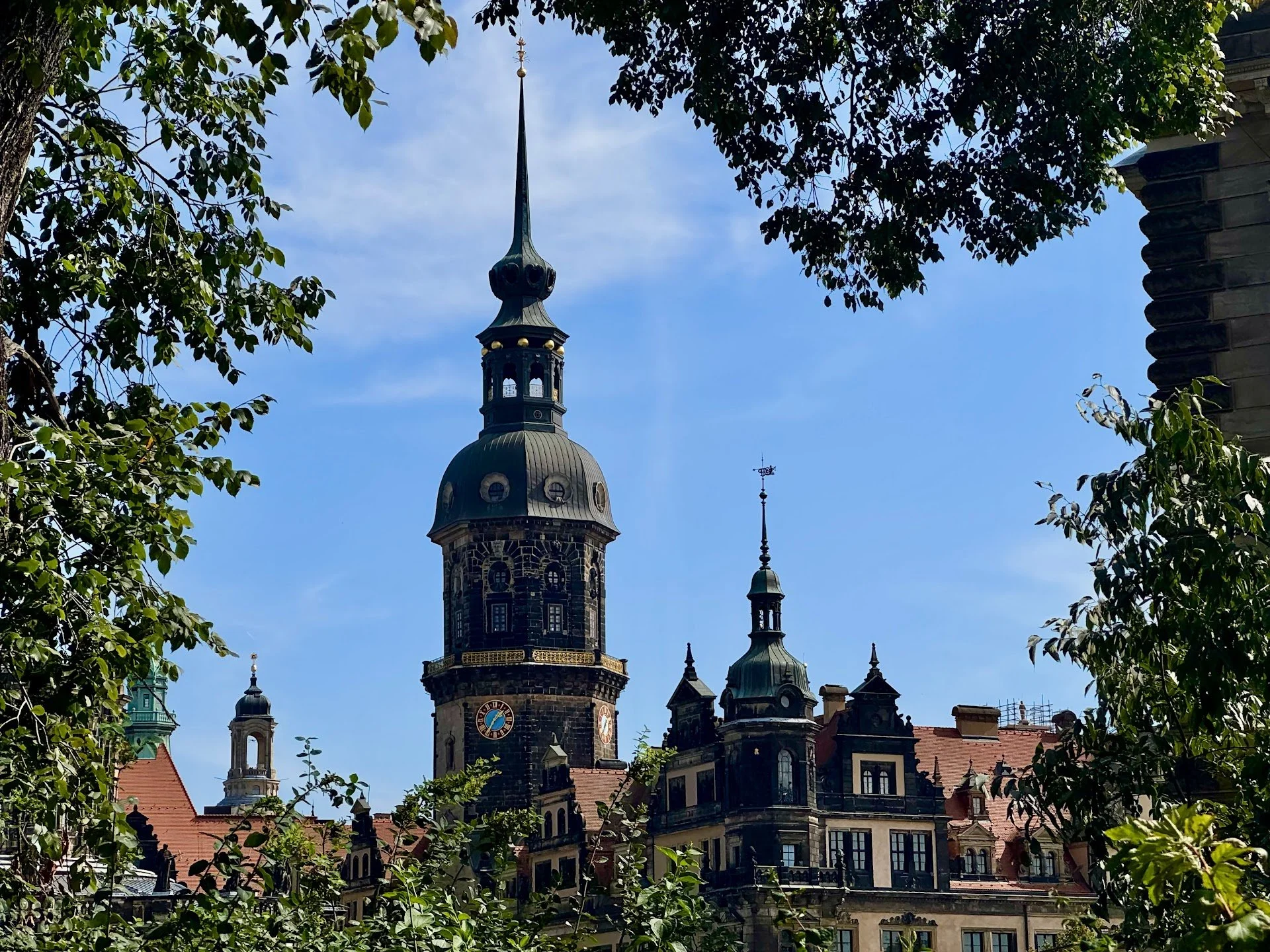 View of a historic building with multiple towers and spires, peeking through green trees against a blue sky.
