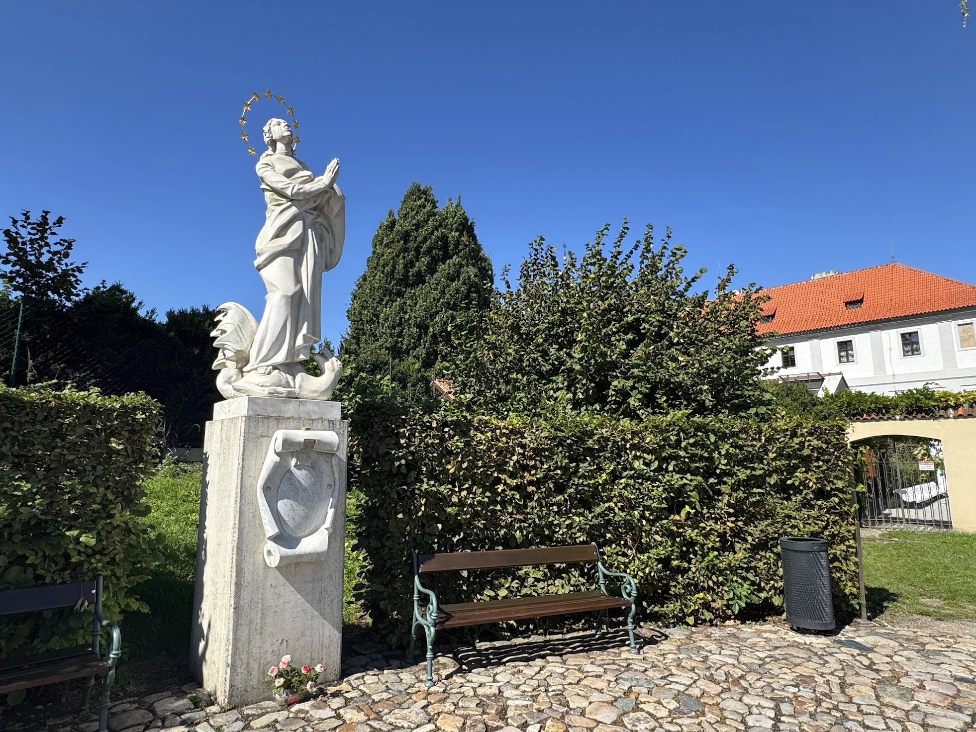 A white religious statue of a woman with hands folded in prayer, standing on a pedestal in a park with green bushes and trees. The background includes a blue sky and buildings with red-tiled roofs.