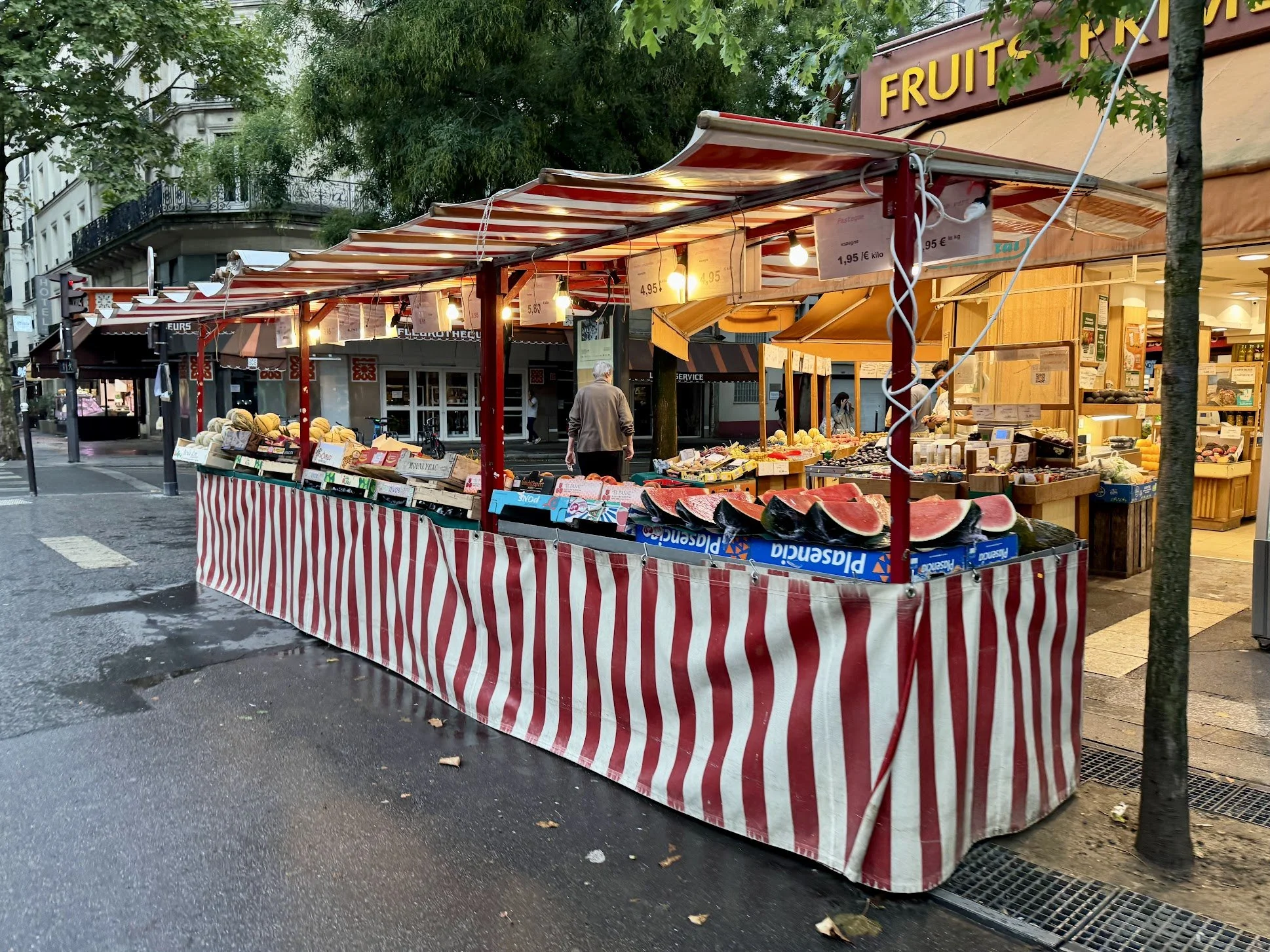 An outdoor fruit market stall with a red and white striped cloth covering the display table. Various cut melons such as watermelons and other fruits are arranged on the tables. Overhead, there are hanging signs with prices for different fruits, and w