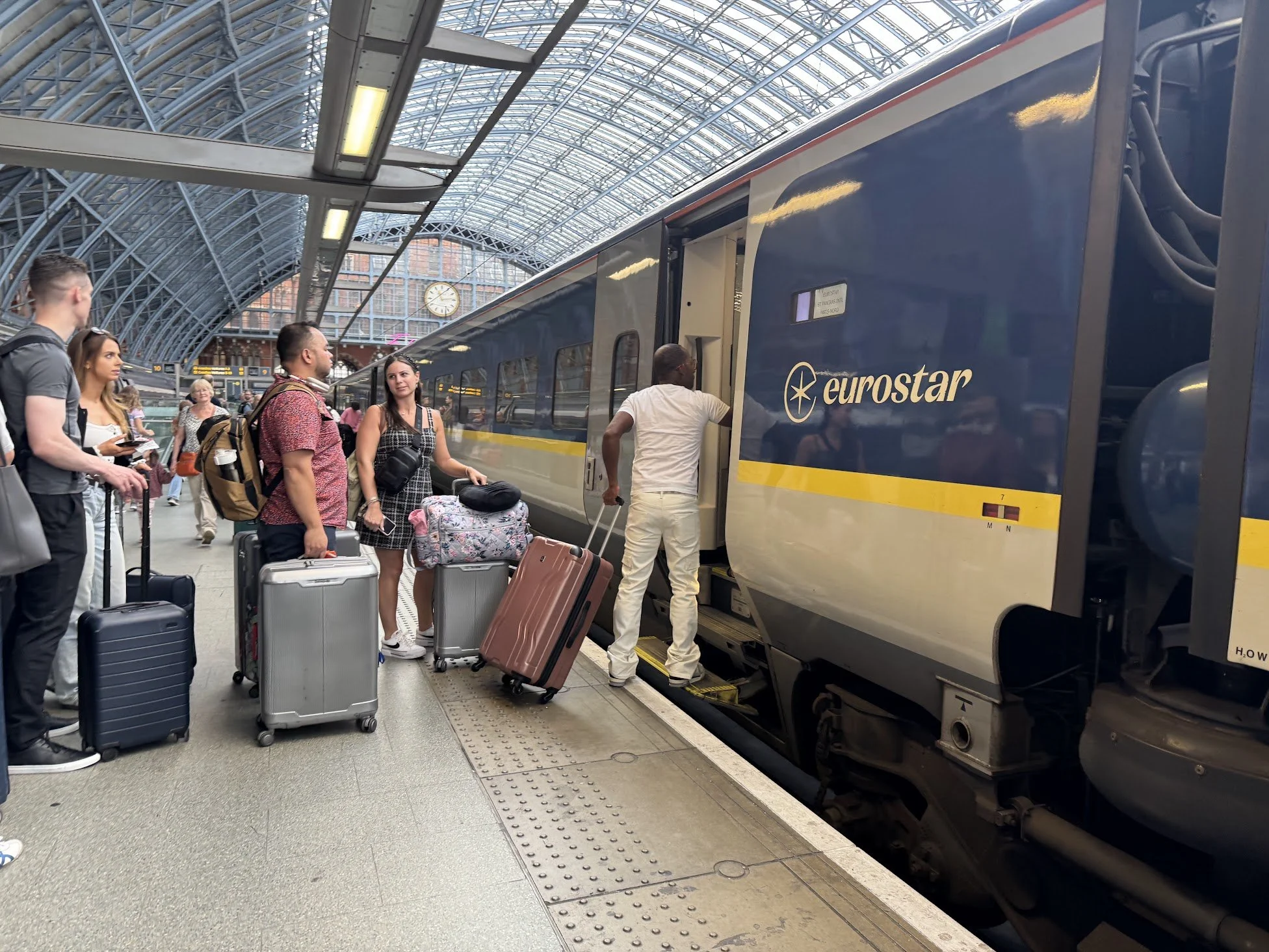 People with luggage boarding a Eurostar train at a station with a large glass and steel arched roof and a clock on the wall in the background.