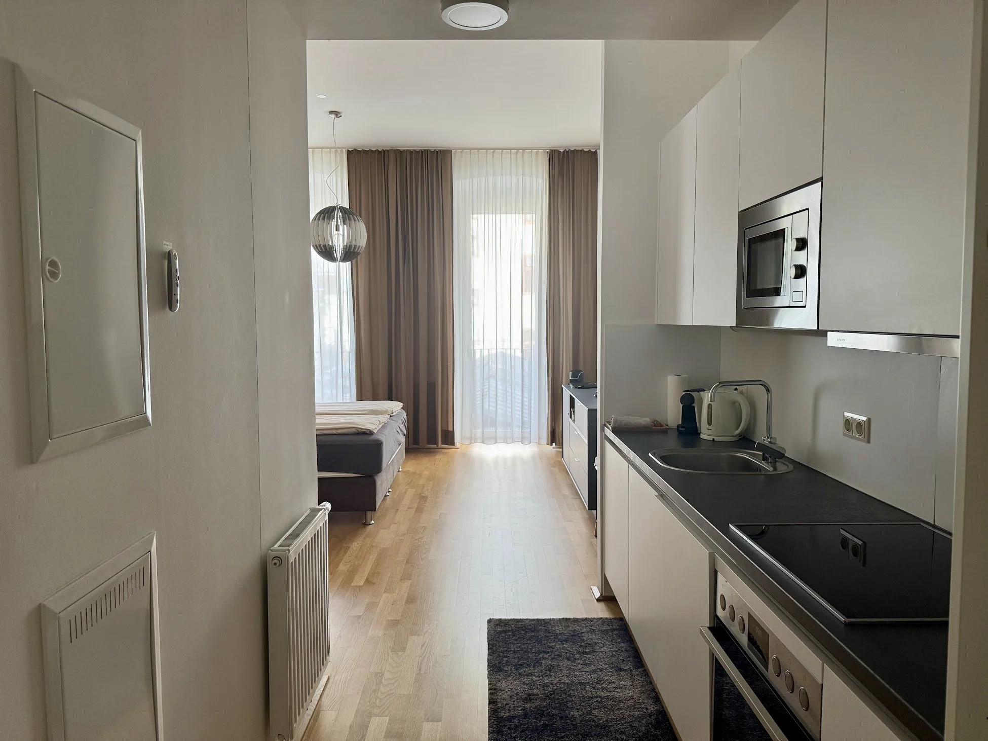 A small studio apartment with an open layout featuring a kitchenette on the right side with white cabinets, a microwave, and a black countertop. In the background, there is a living area with a bed, large windows with sheer and brown curtains, and a 