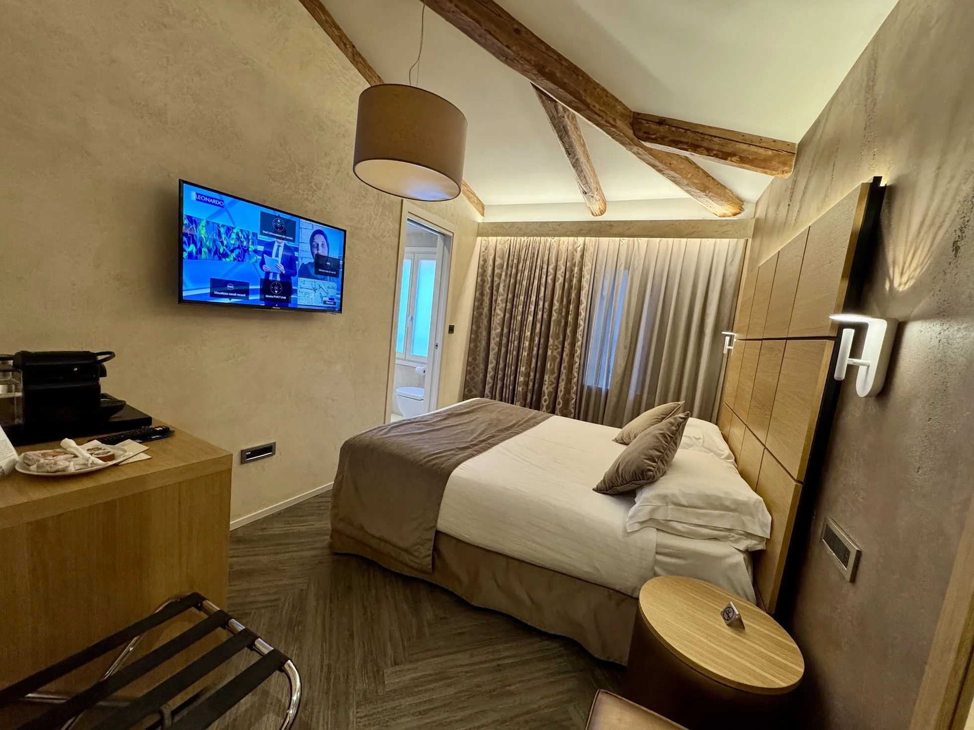 A hotel room with a bed, wooden ceiling beams, a wall-mounted TV playing a show, curtains covering a window, and a small bedside table with a lamp.
