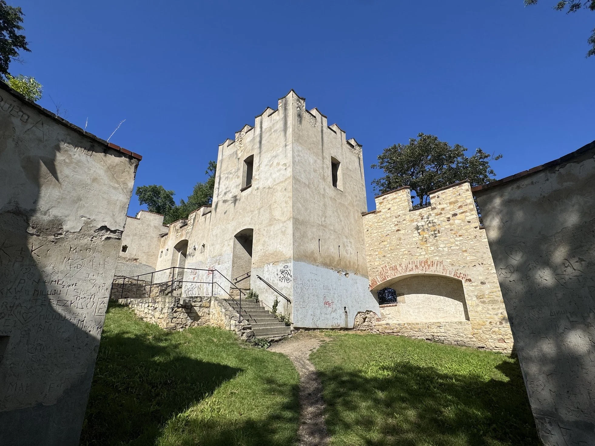 A historic stone building with crenellated walls and tall narrow windows, surrounded by graffiti-covered walls and green grass, with trees and a bright blue sky in the background.
