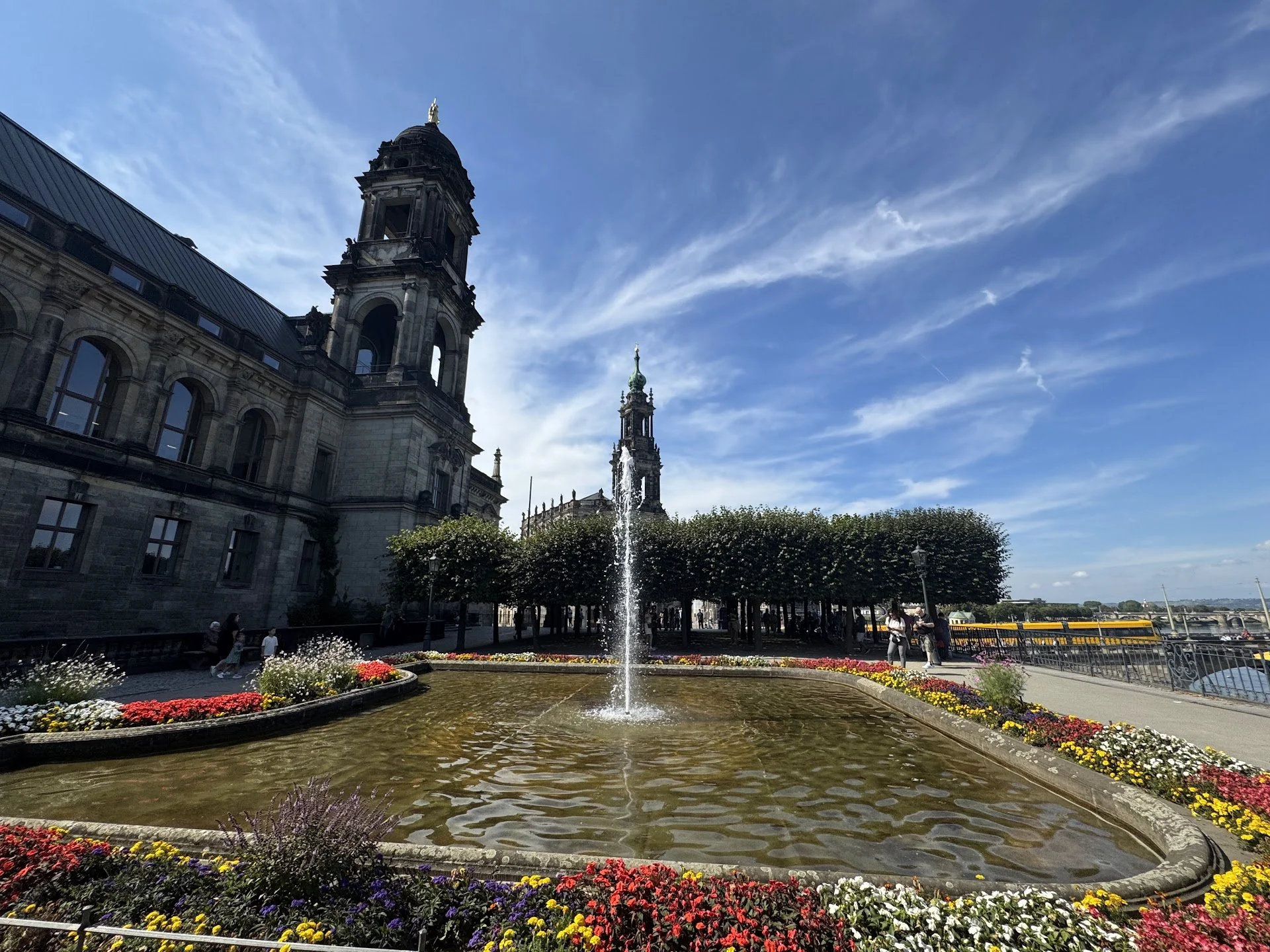 View of historic building with clock towers, fountain in front, and flower garden, under a blue sky with wispy clouds.