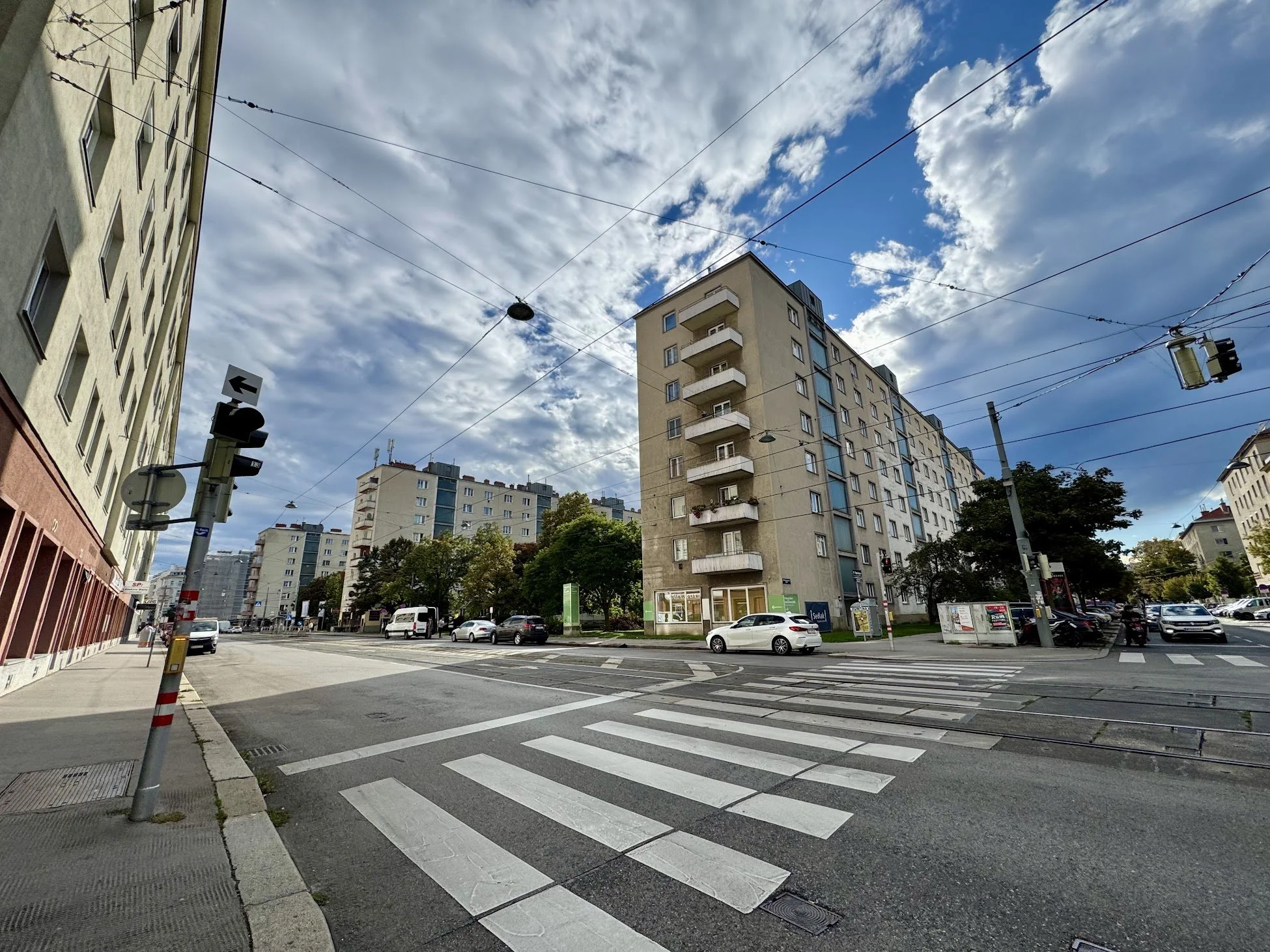 City street with cars, buildings, traffic lights, and a crosswalk under a partly cloudy sky.