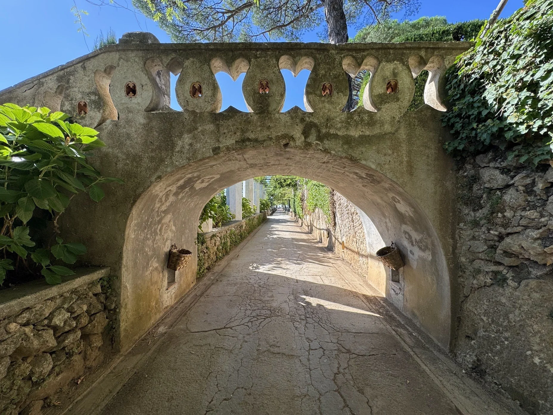 Stone archway with a pathway, surrounded by greenery and a clear blue sky.