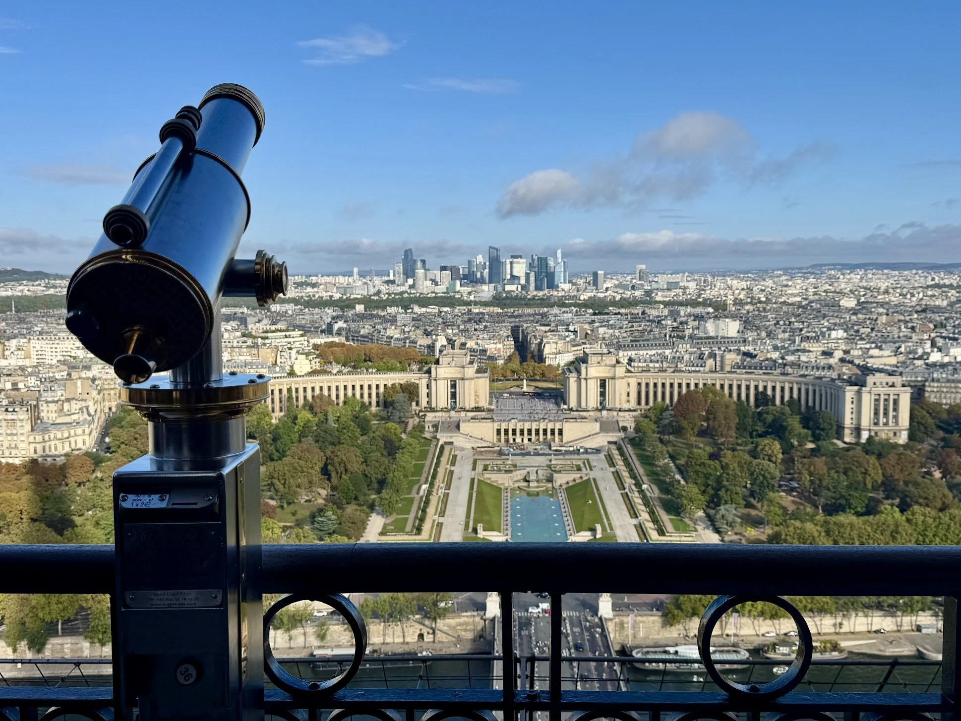 A view of a city skyline with tall modern skyscrapers in the distance, taken from a high vantage point, with a telescope in the foreground.