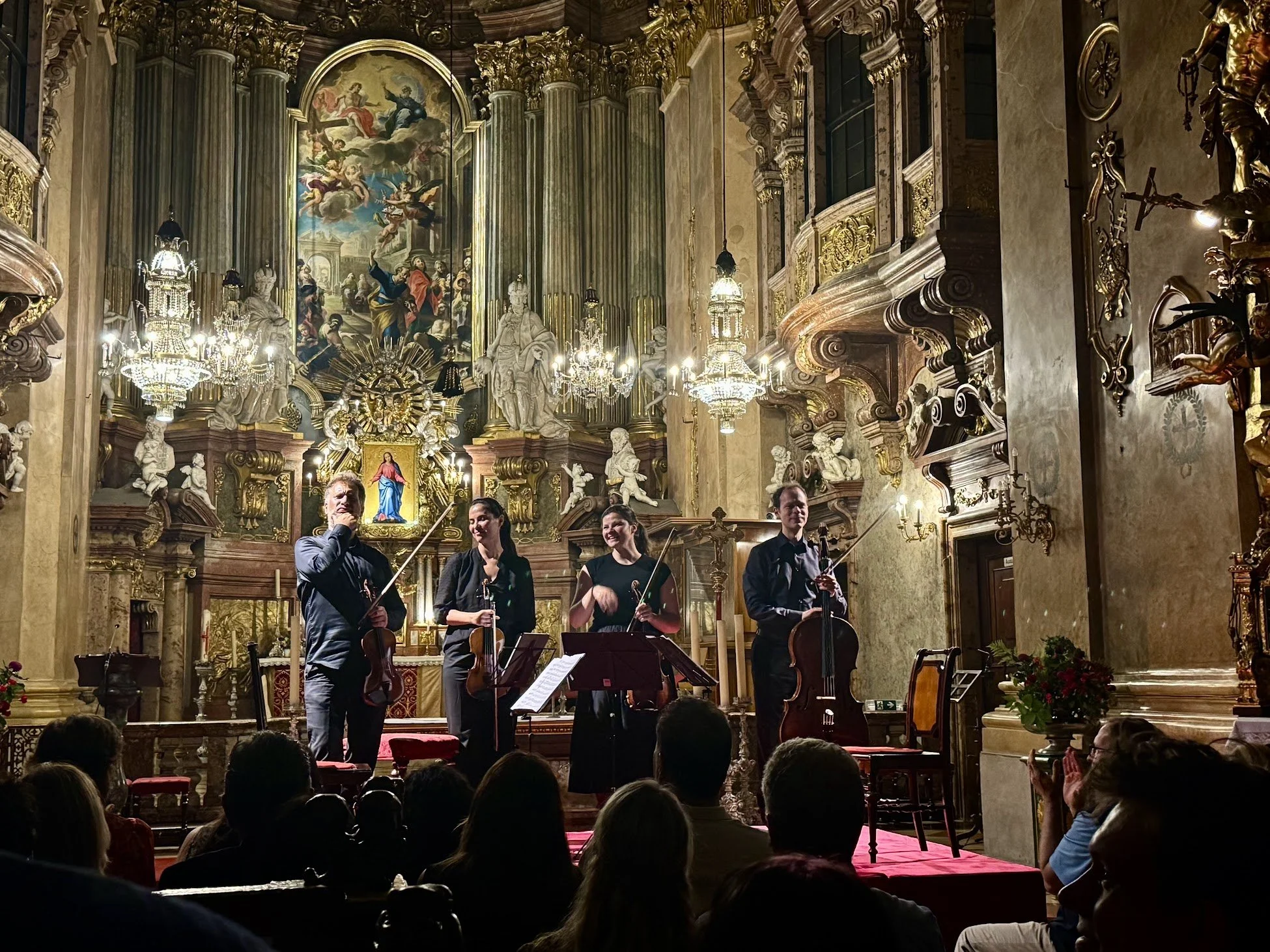 A group of four musicians playing string instruments during a classical concert inside an ornate church or cathedral decorated with gold accents, statues, chandeliers, and religious artwork.