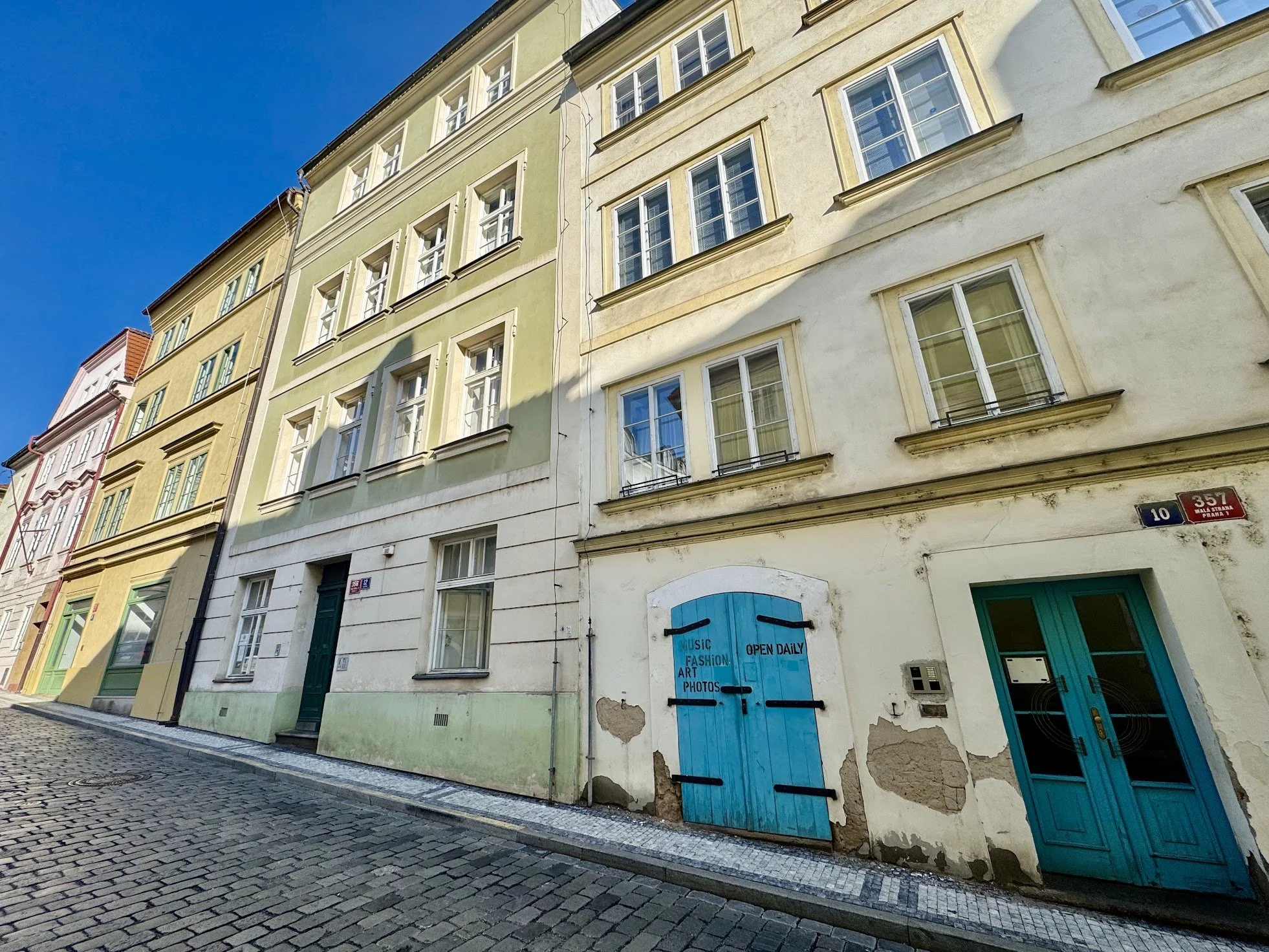 Colorful old buildings with green and beige facades on a cobblestone street under a clear blue sky. Some windows are open and a door with blue paint has city signs and a small window.