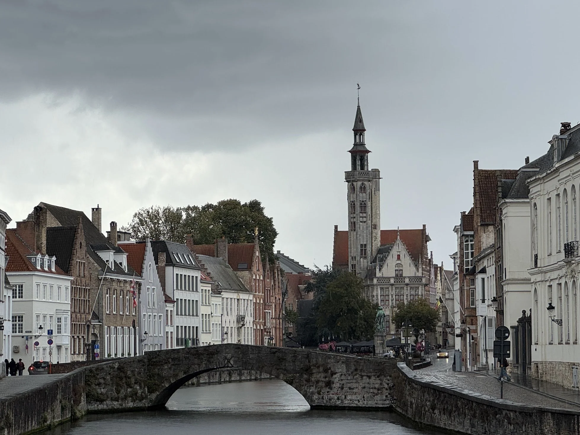 A cityscape with old European-style buildings along a canal, a stone bridge crossing over the canal, and a church tower under a cloudy sky.