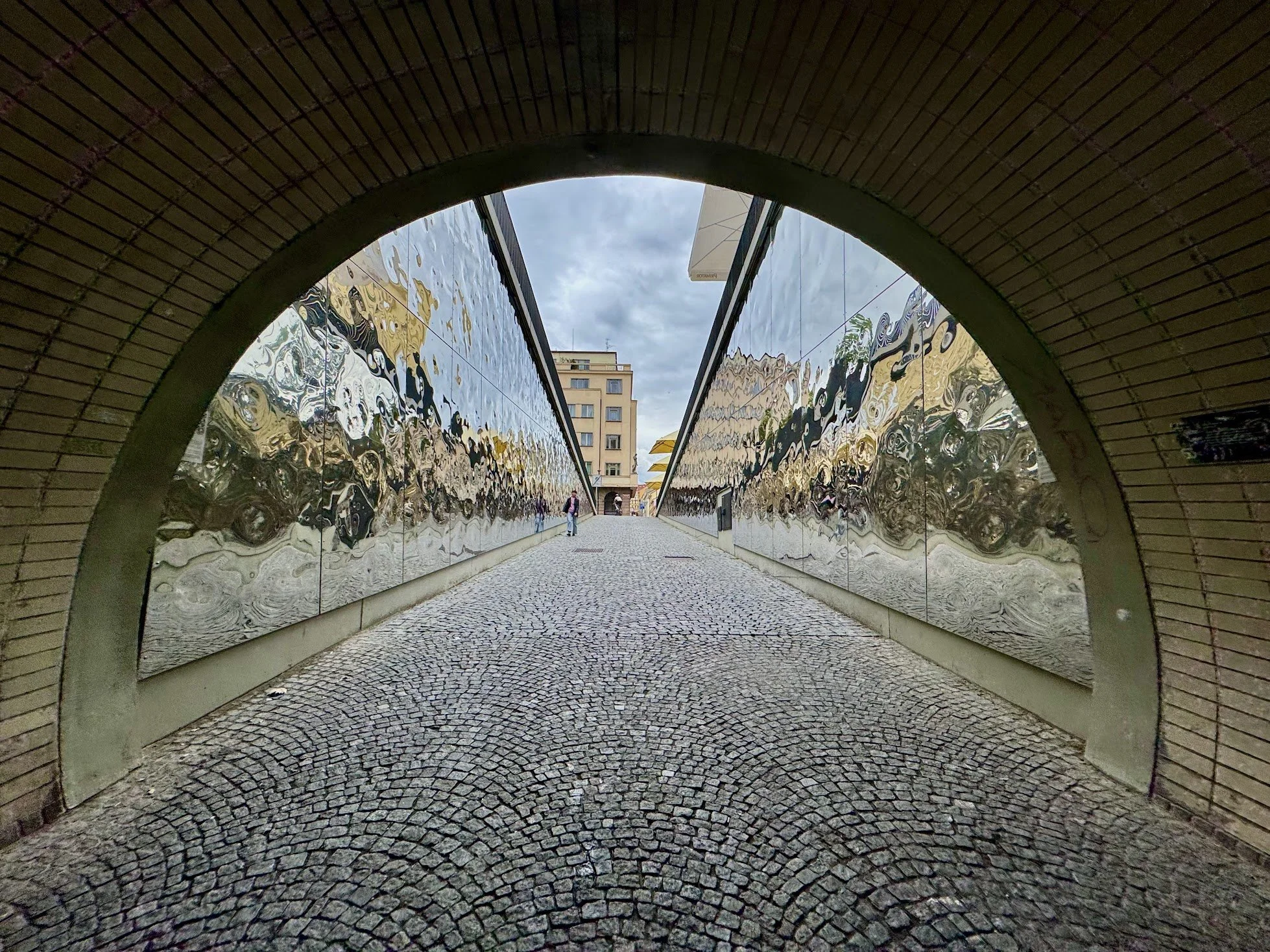 Image taken from underneath an arched tunnel with brick walls, looking out into a city street with a building, cloudy sky, and a person walking.