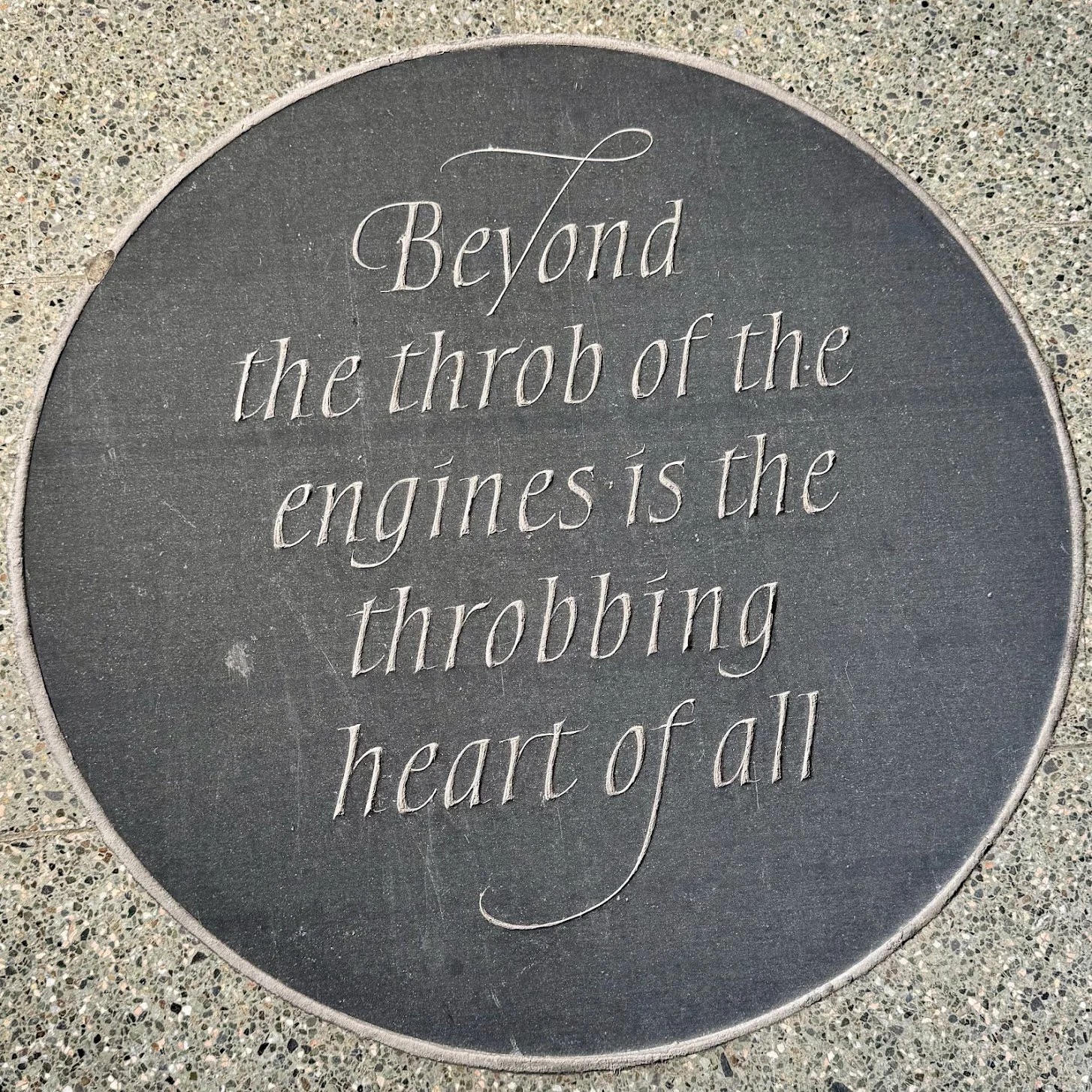 A circular plaque with an inspirational quote engraved on it, set into a speckled concrete surface. The quote reads: 'Beyond the throbbing of the engines is the throbbing heart of all.'