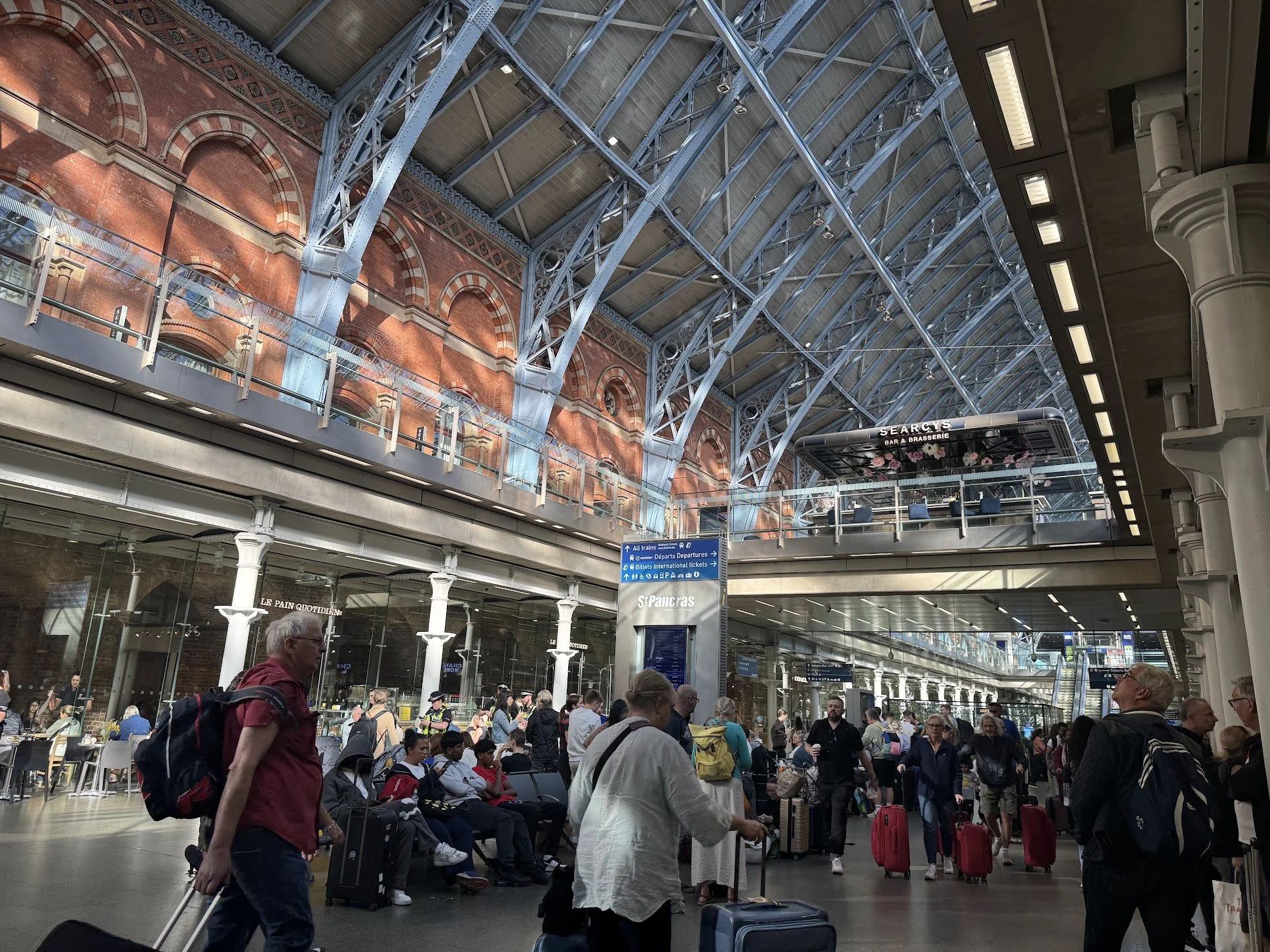 People with luggage waiting and walking inside a busy train station with high arched glass and metal ceiling, brick wall, and signage indicating train departures and international tickets.