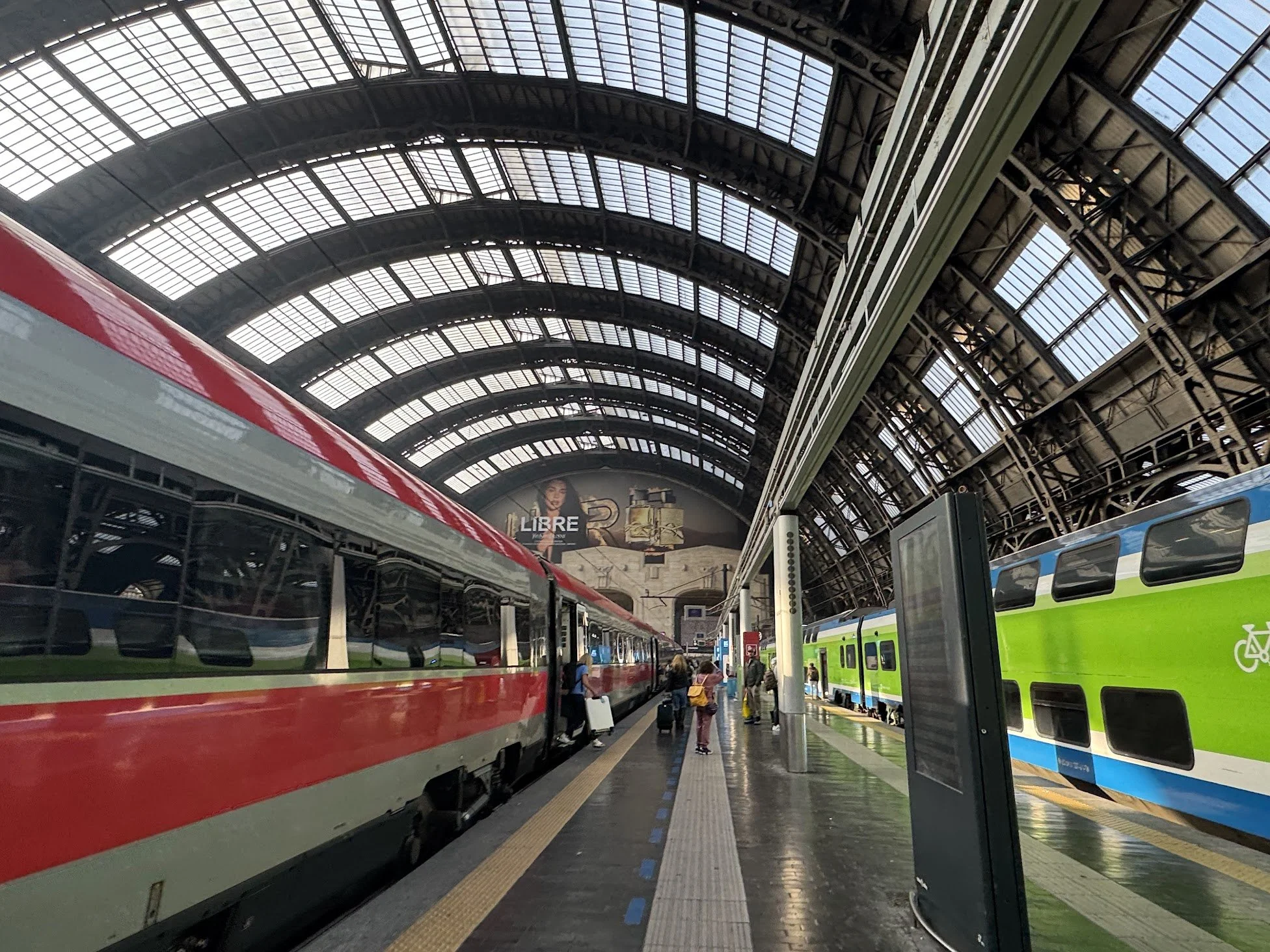 Inside a large train station with arched glass roof, multiple trains on tracks, including a red train on the left and a green and blue train on the right, with passengers walking and waiting.