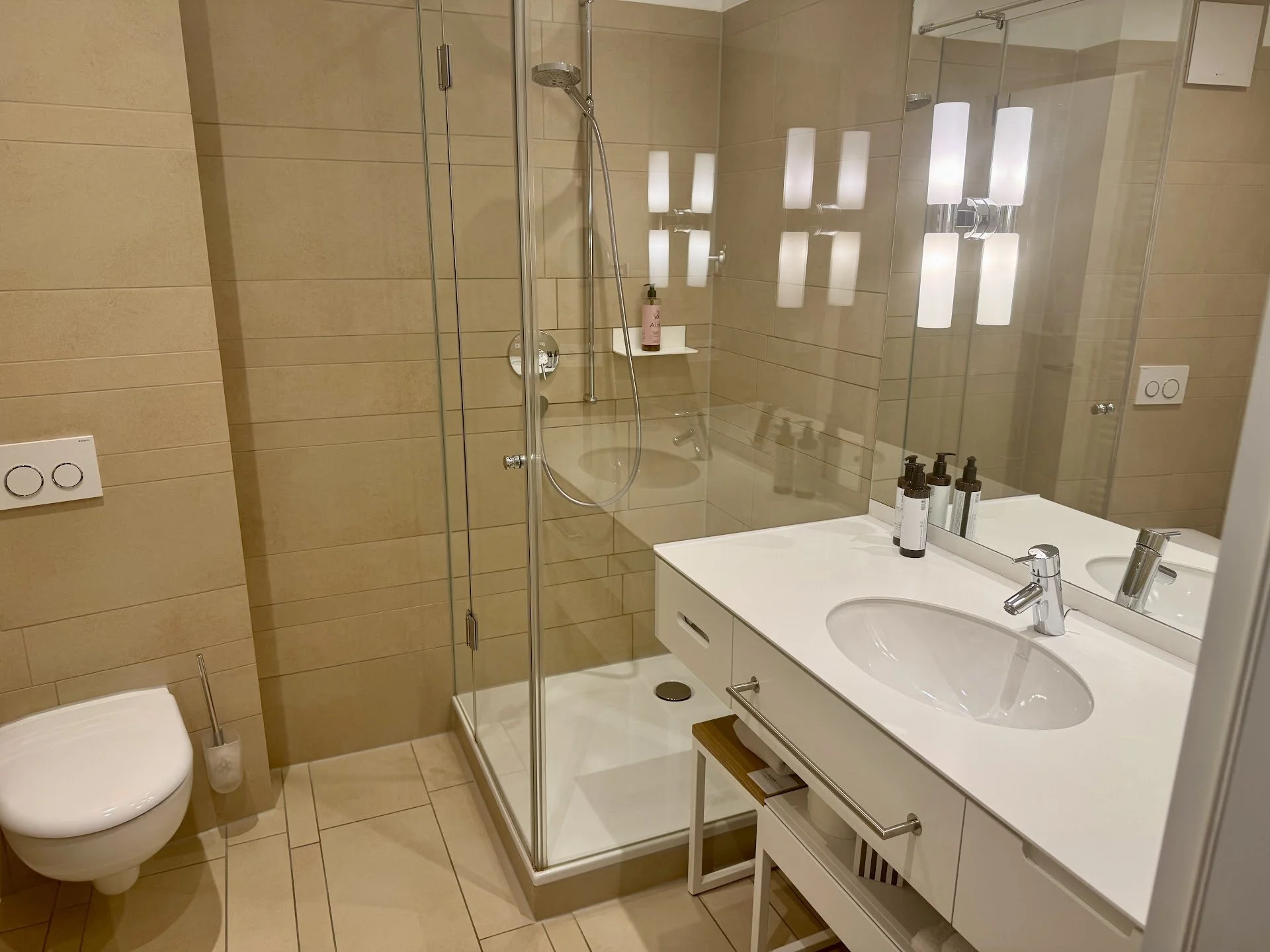 Modern bathroom with beige tiled walls and floor, a white toilet, a glass-enclosed shower with a handheld showerhead, a white vanity with two sinks, a large mirror, and contemporary light fixtures. There are three bottles of toiletries on the vanity 