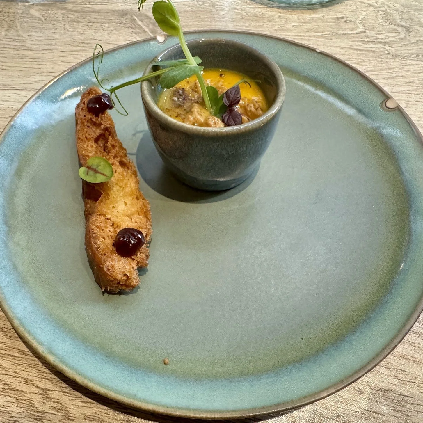A blue ceramic plate with a small cup of baked eggs garnished with microgreens and herbs, and a long toasted breadstick topped with cherries and microgreens.
