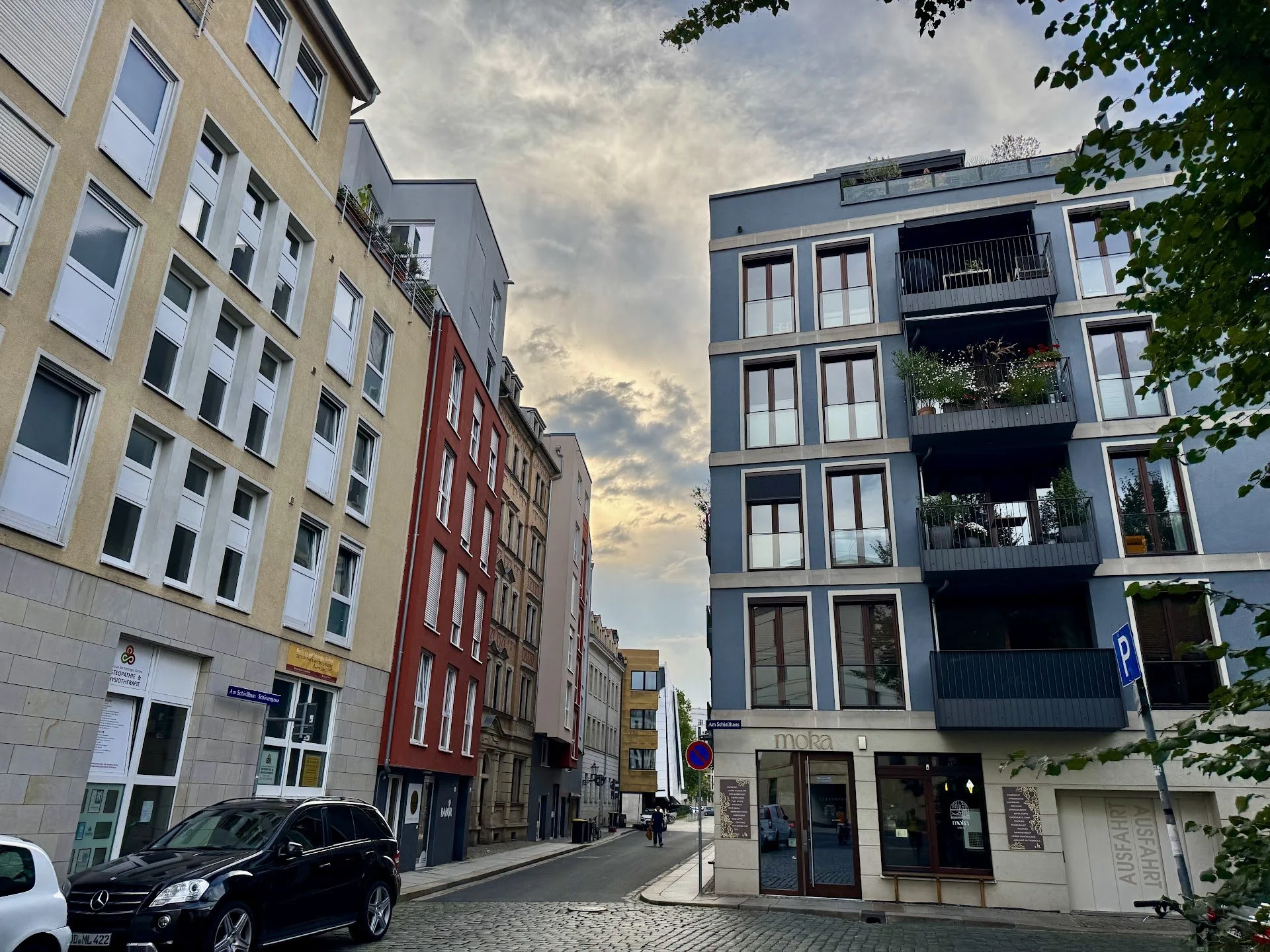 View of a city street with modern multi-story buildings, parked cars, a person walking, and a cloudy sky overhead.