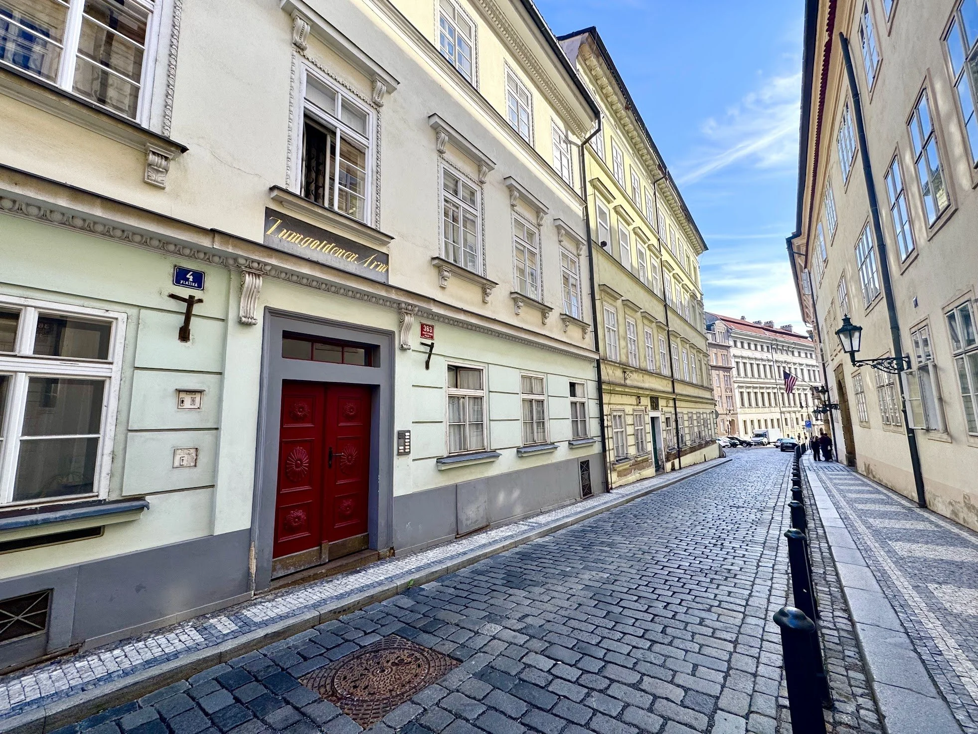 A cobblestone street lined with historic European-style buildings with ornate window frames and decorative details. There is a red door and a street sign with the number 4 and the street name in a foreign language. The sky is clear and blue, suggesti