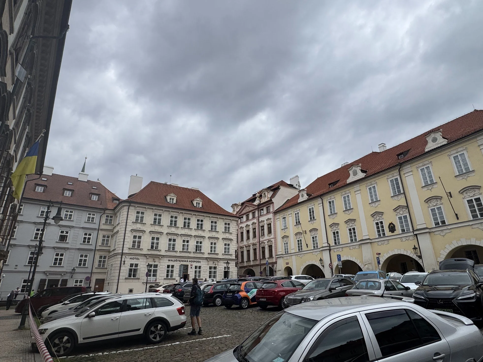 A city square with historic buildings, a parking lot filled with cars, and overcast cloudy sky. There is a yellow and blue flag on a pole on the left side.