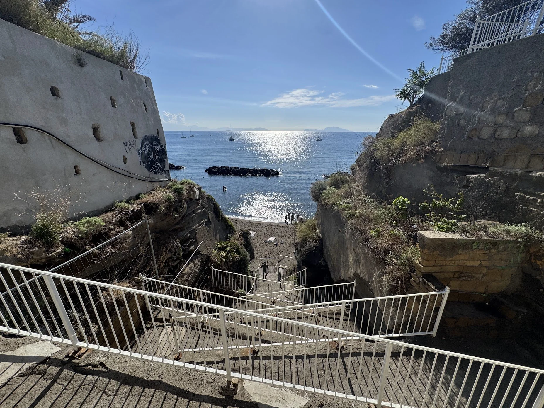 A stairway leading down to a beach with rocks and umbrellas, framed by a white fence, with the ocean and sailboats in the background under a sunny sky.