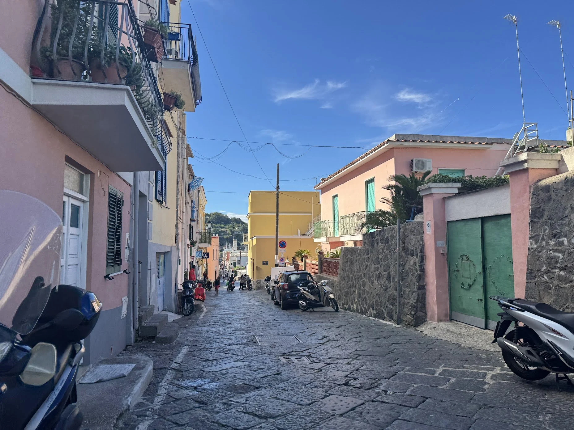 A narrow cobblestone street lined with colorful buildings and parked motorcycles and cars. People are walking and the sky is clear with a few clouds.